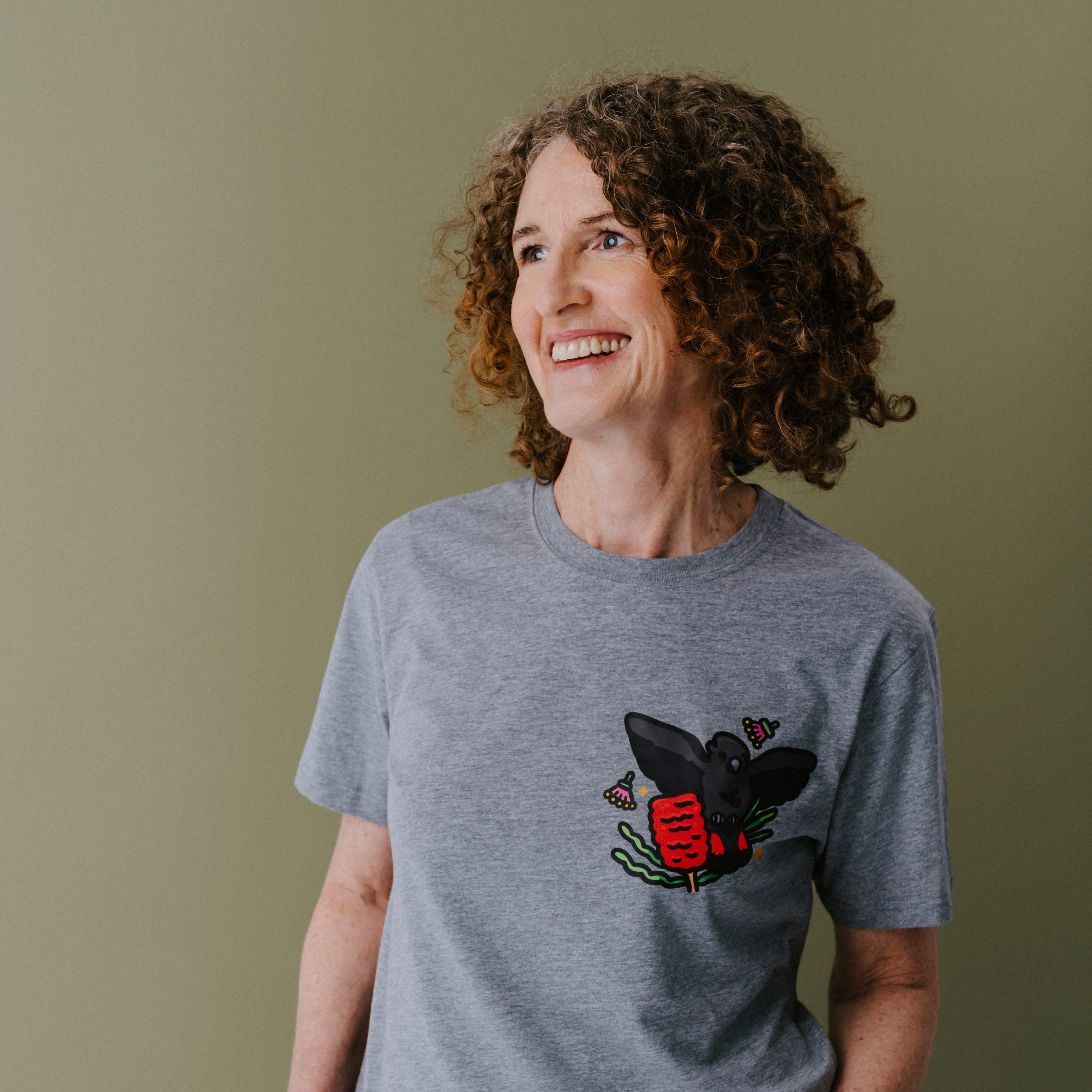 A smiling woman with curly brown hair wearing a heather gray t-shirt with a glossy black cockato illustration on the chest, set against a solid olive green background.