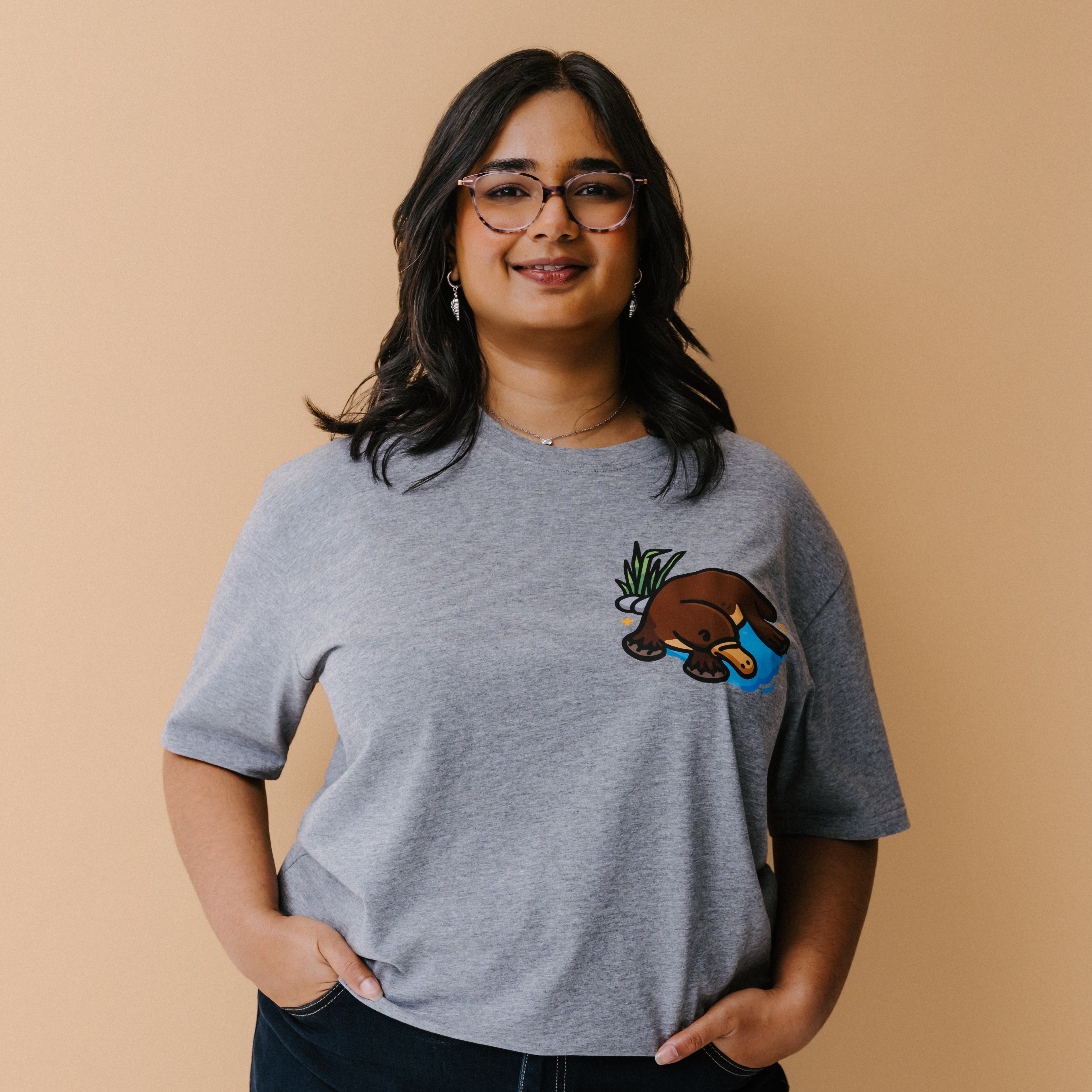 A woman with glasses is wearing a heather gray t-shirt with a small illustrated platypus in a blue pond and green plant on the chest, set against a solid beige background.