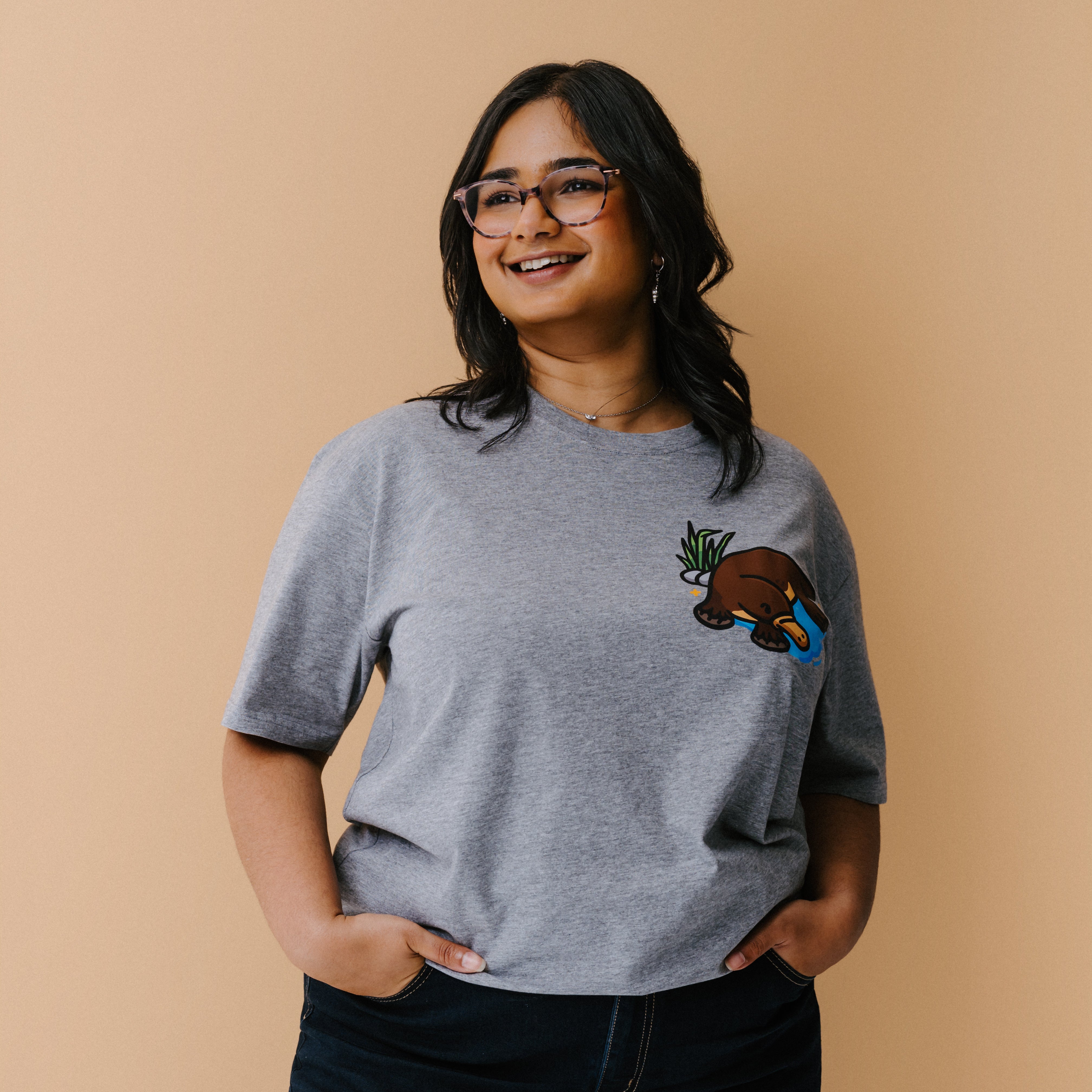 A woman with glasses is wearing a heather gray t-shirt with a small illustrated platypus in a blue pond and green plant on the chest, set against a solid beige background.