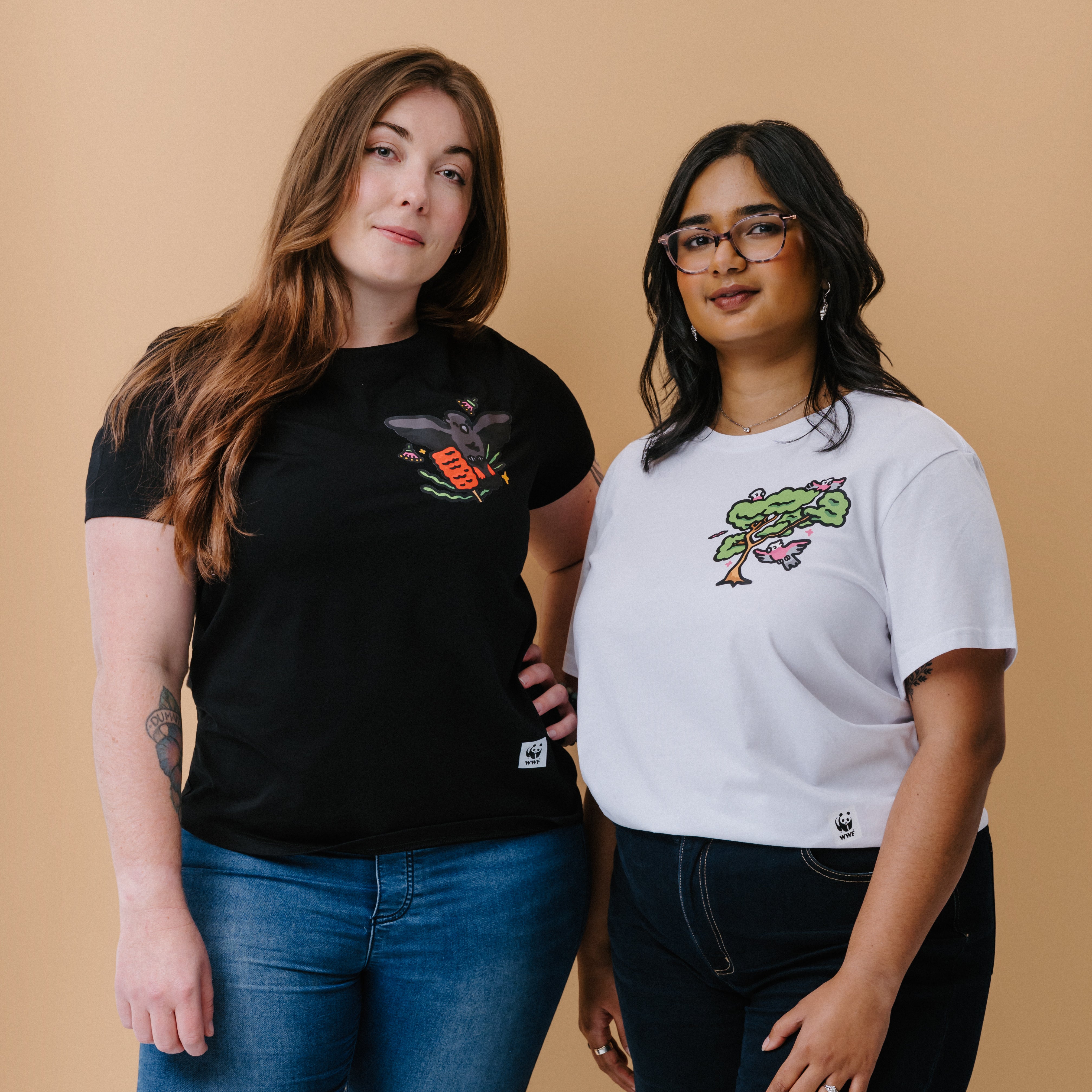 Two women modeling graphic t-shirts against a solid beige background. The woman on the left wears a black crewneck t-shirt with a glossy black cockatoo graphic, and the woman on the right wears a white crewneck t-shirt with a green tree graphic.