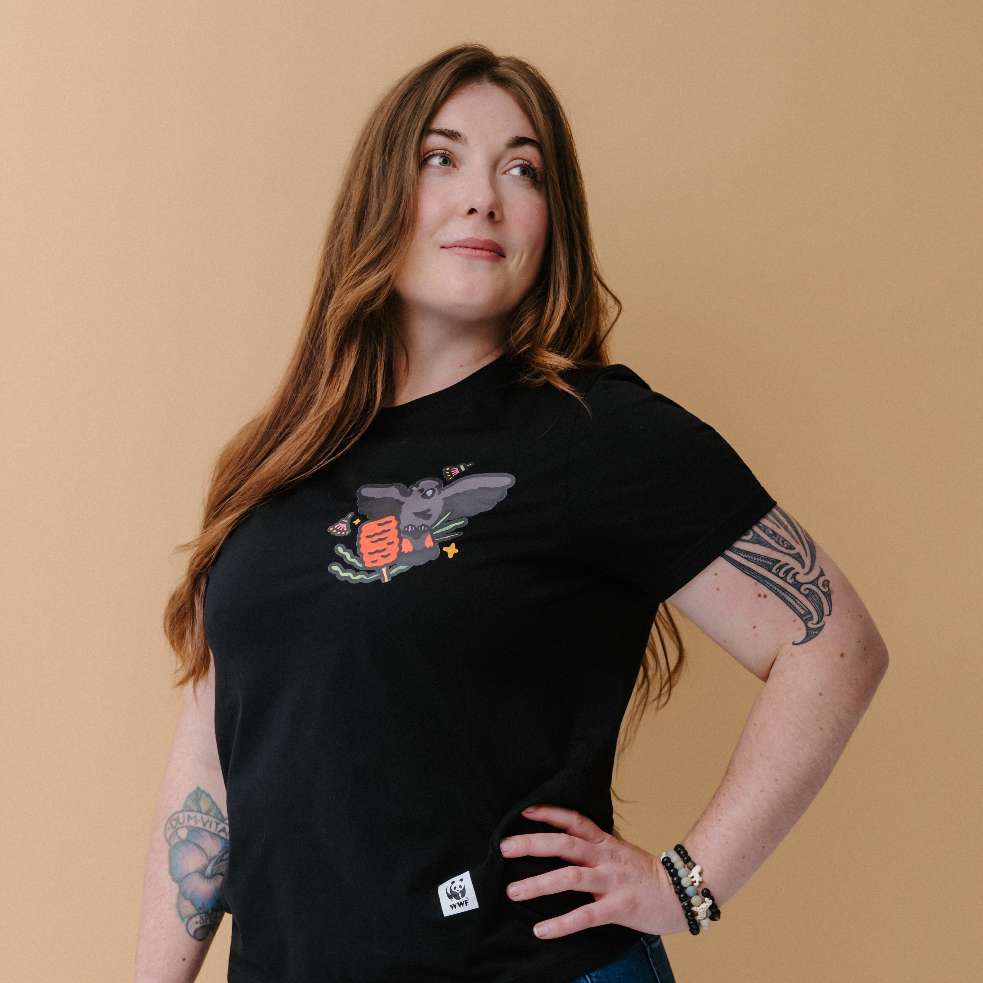 A woman with long brown hair and tattoos wearing a black graphic t-shirt with an illustrated glossy black cockatoo, posing against a solid beige background.
