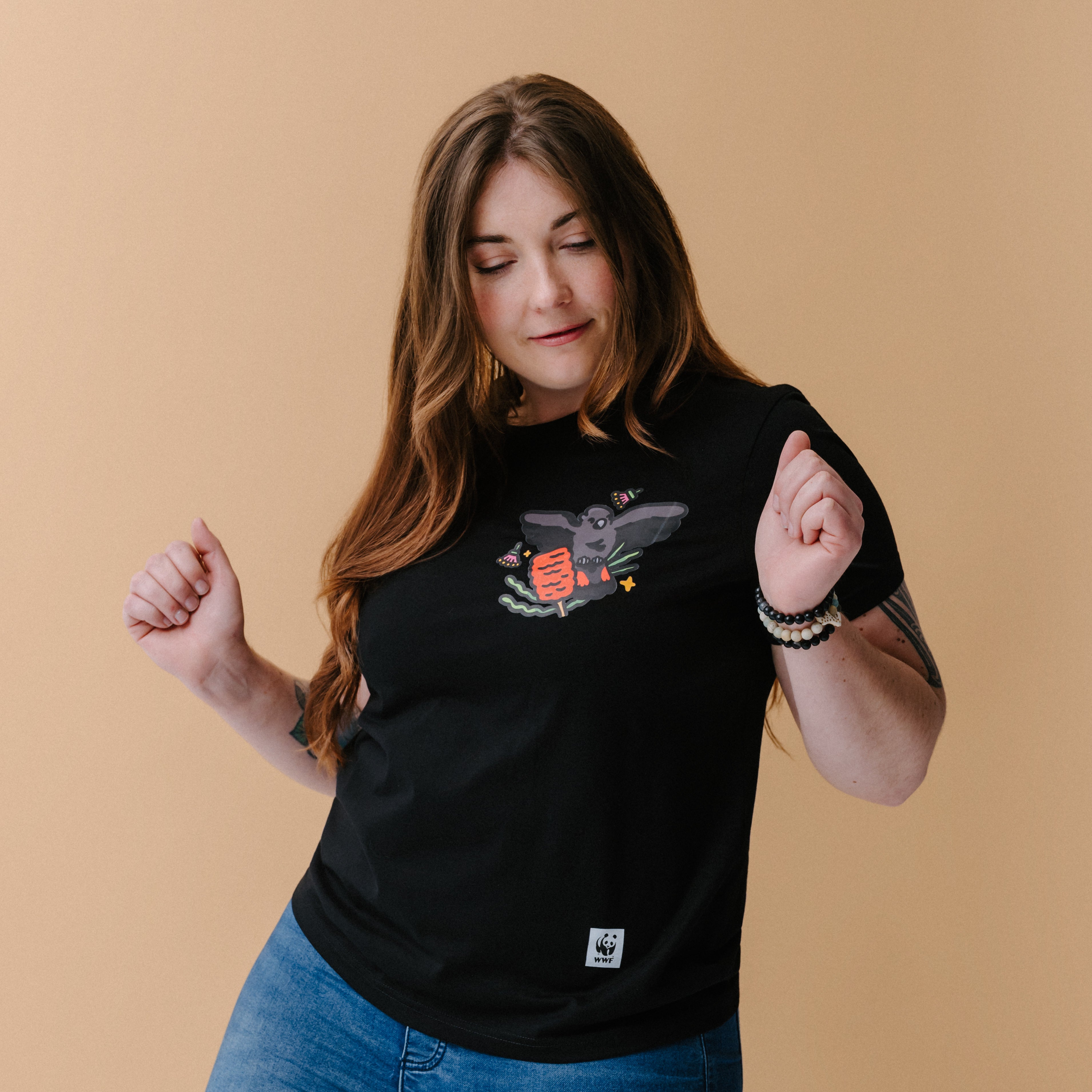 A woman with long brown hair and tattoos wearing a black graphic t-shirt with an illustrated glossy black cockatoo, posing against a solid beige background.