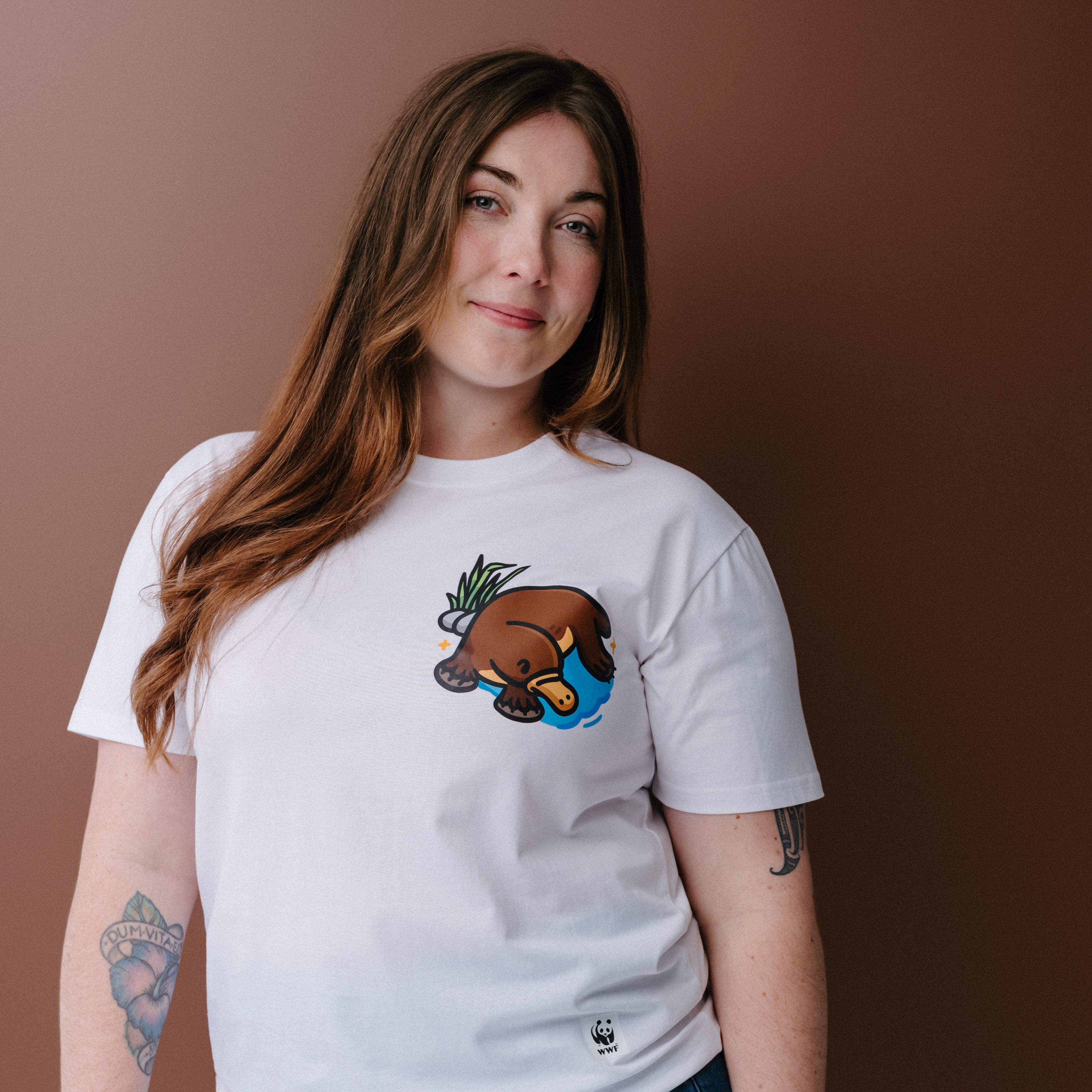 A woman with long brown hair wearing a white crewneck t-shirt with an illustrated platypus graphic on the chest, posed against a solid brown background.