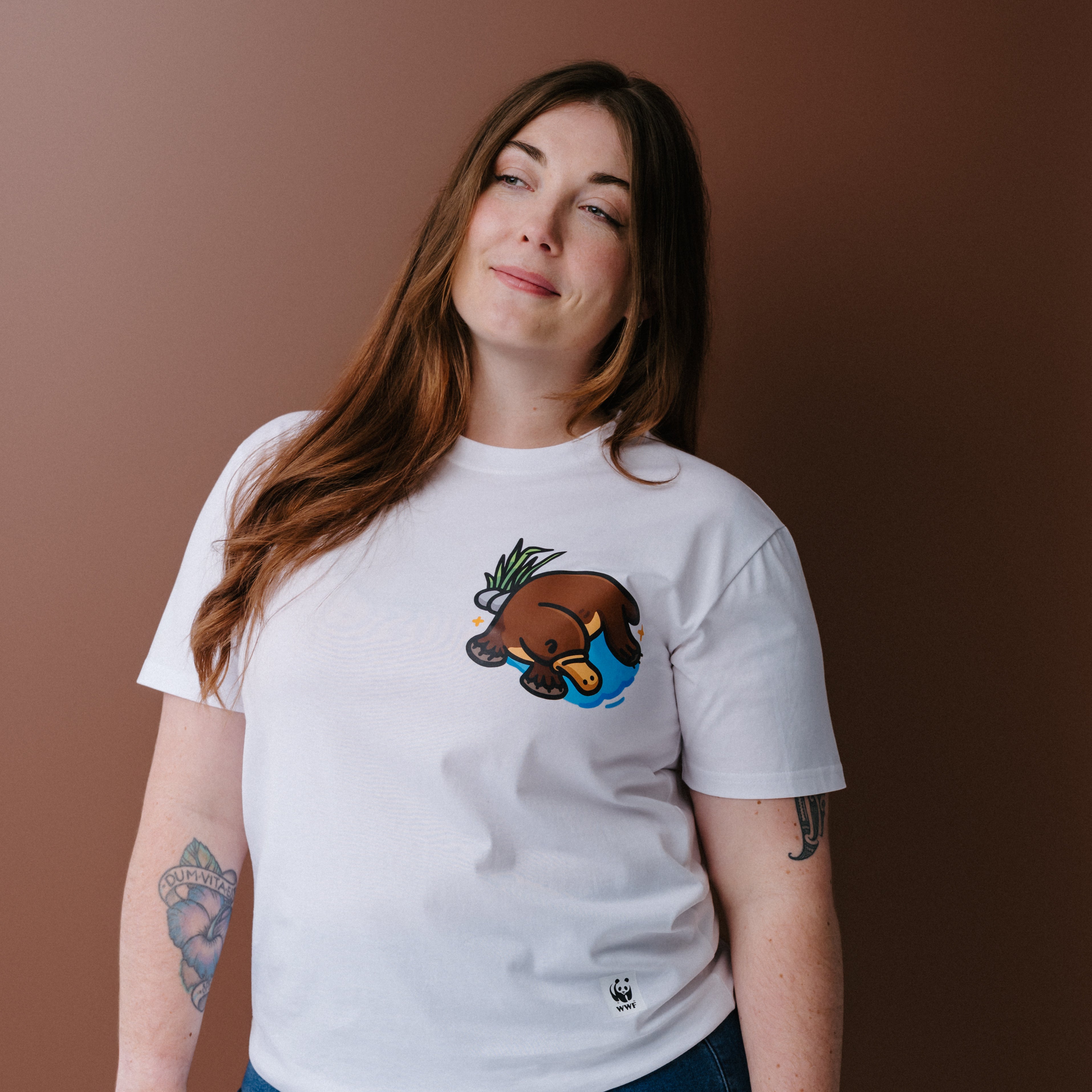 A woman with long brown hair wearing a white crewneck t-shirt with an illustrated platypus graphic on the chest, posed against a solid brown background.