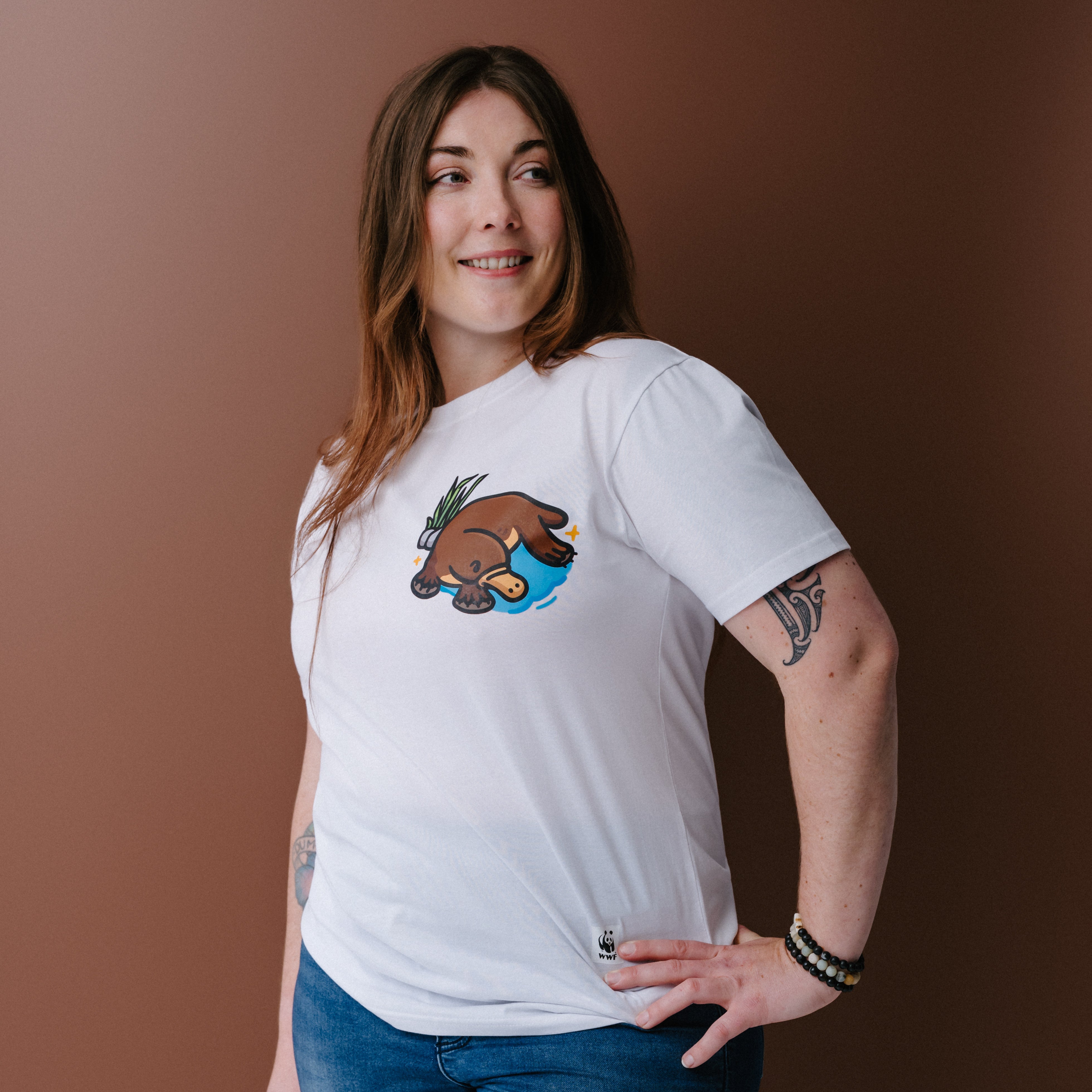 A woman with long brown hair wearing a white crewneck t-shirt with an illustrated platypus graphic on the chest, posed against a solid brown background.