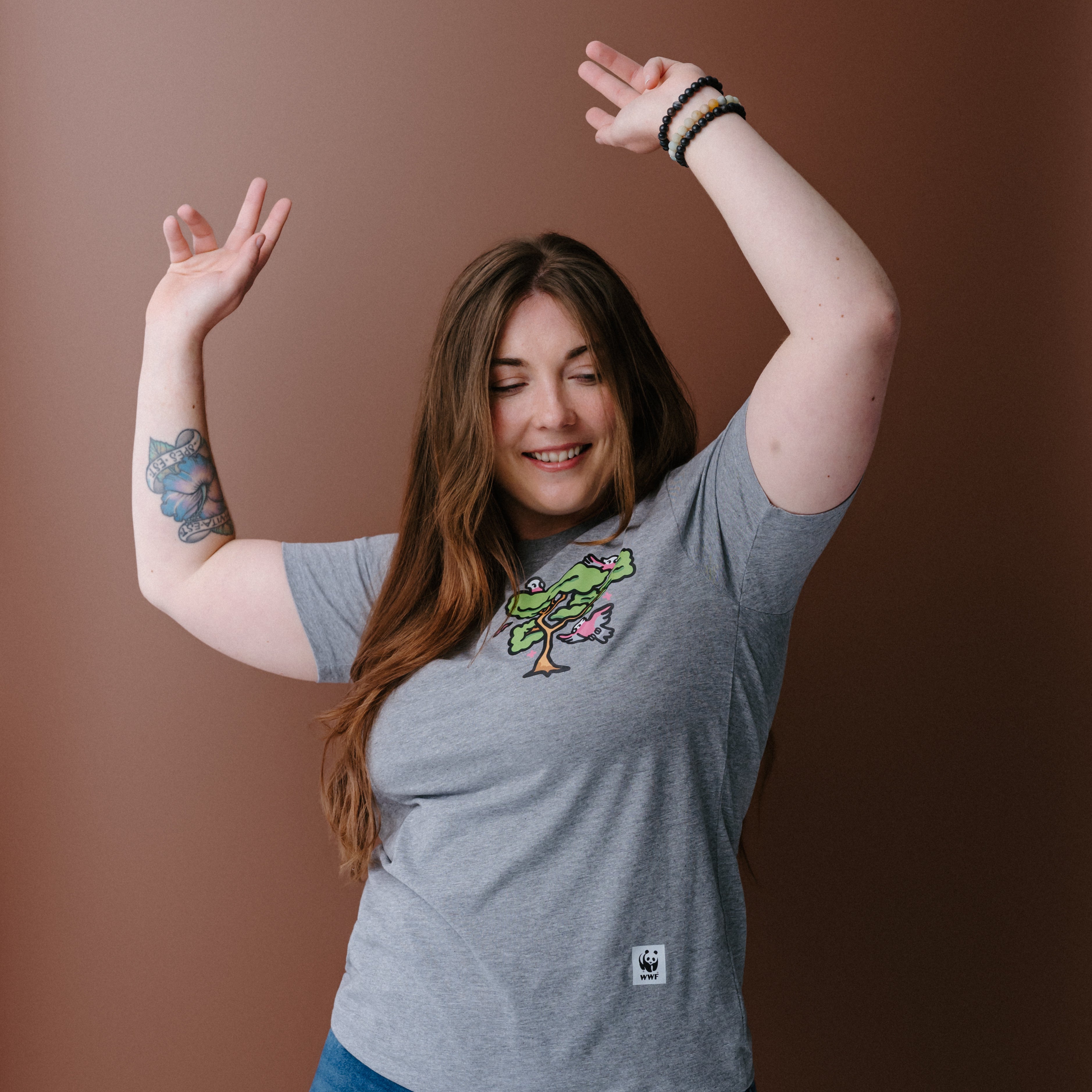 A woman with long brown hair wearing a heather gray t-shirt with a colorful tree and galas illustration on the left chest, posing against a solid brown background.