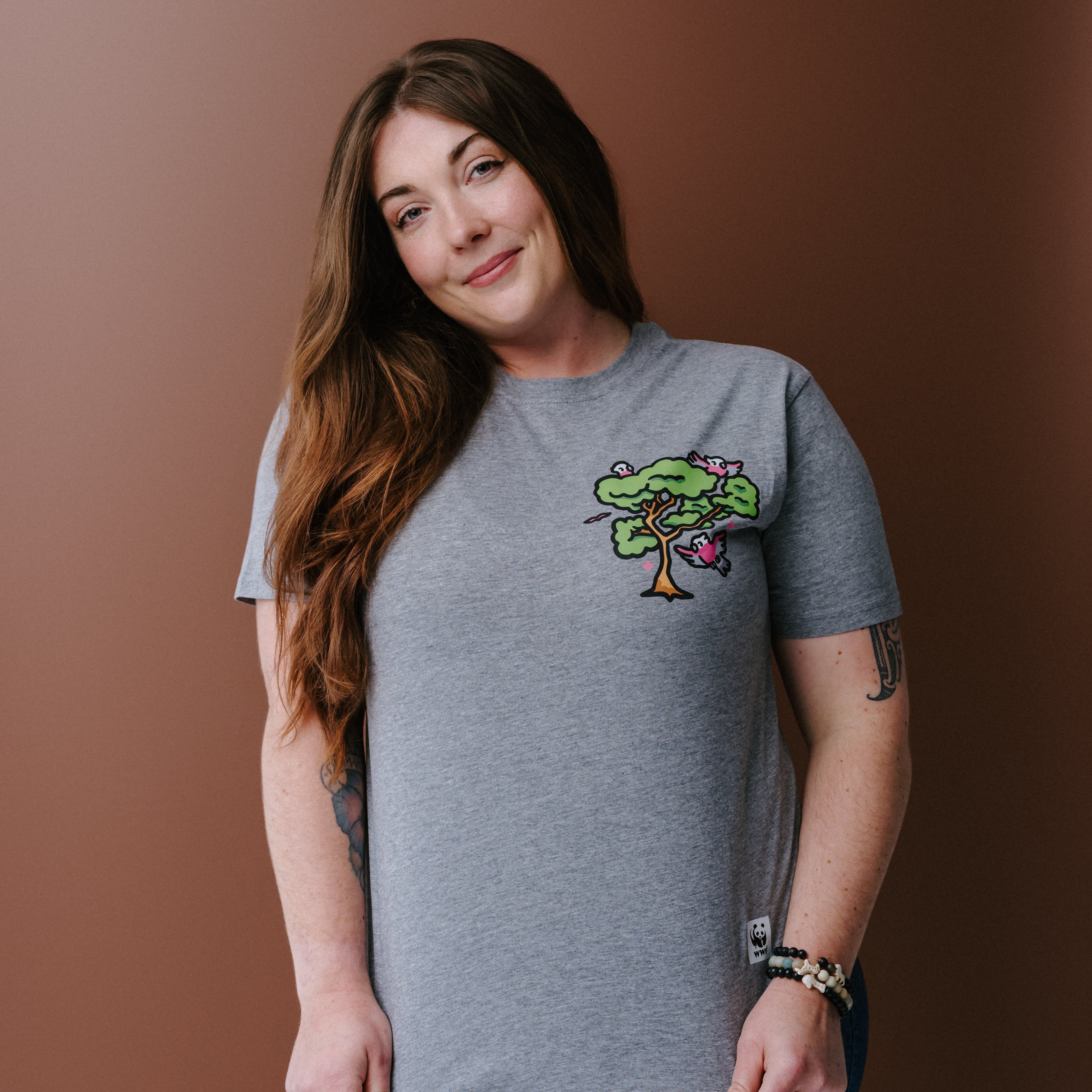 A woman with long brown hair wearing a heather gray t-shirt with a colorful tree and galas illustration on the left chest, posing against a solid brown background.