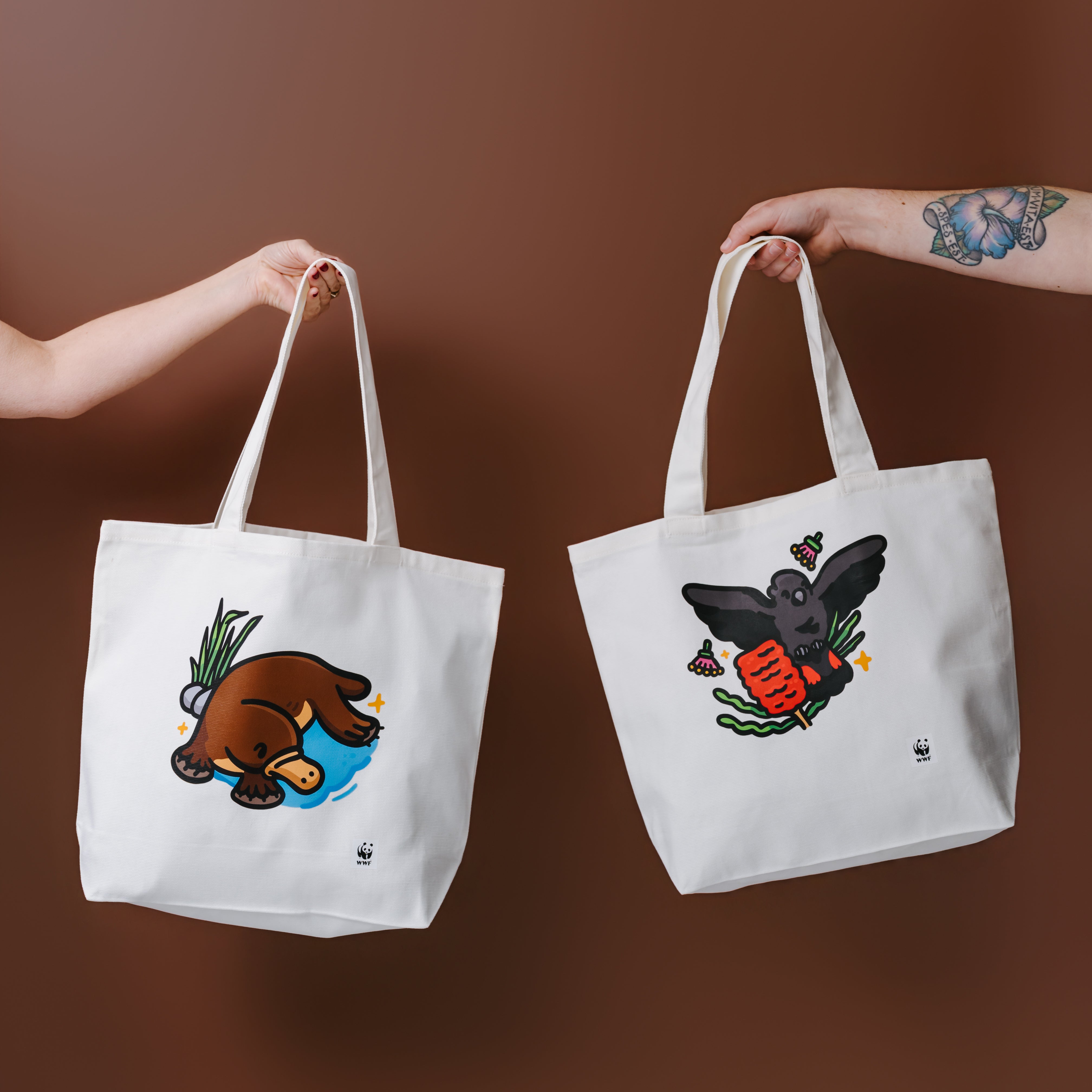 A white canvas tote bag with a graphic of a glossy black cockatoo, held by a person's tattooed arm against a solid brown background.