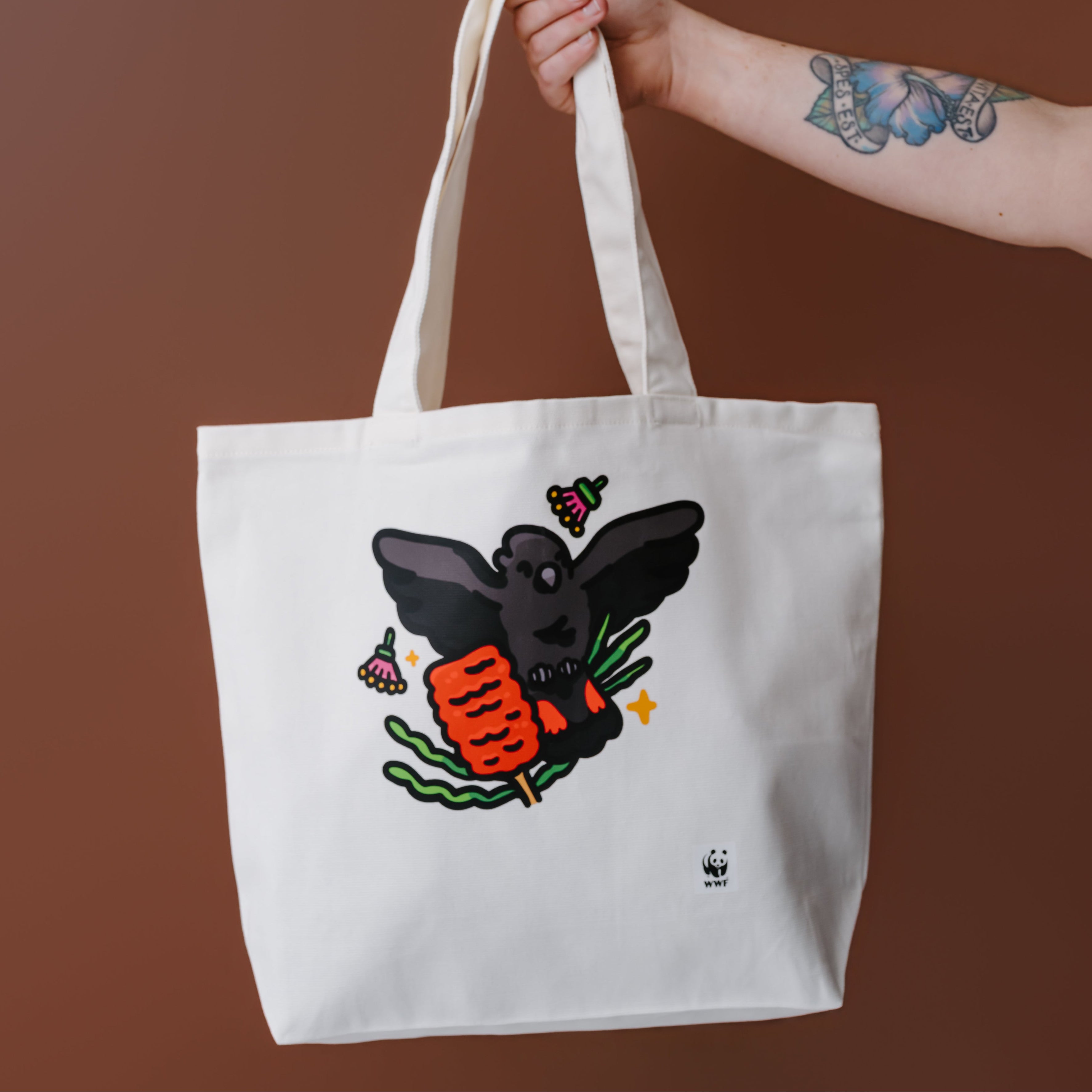 A white canvas tote bag with a graphic of a glossy black cockatoo, held by a person's tattooed arm against a solid brown background.