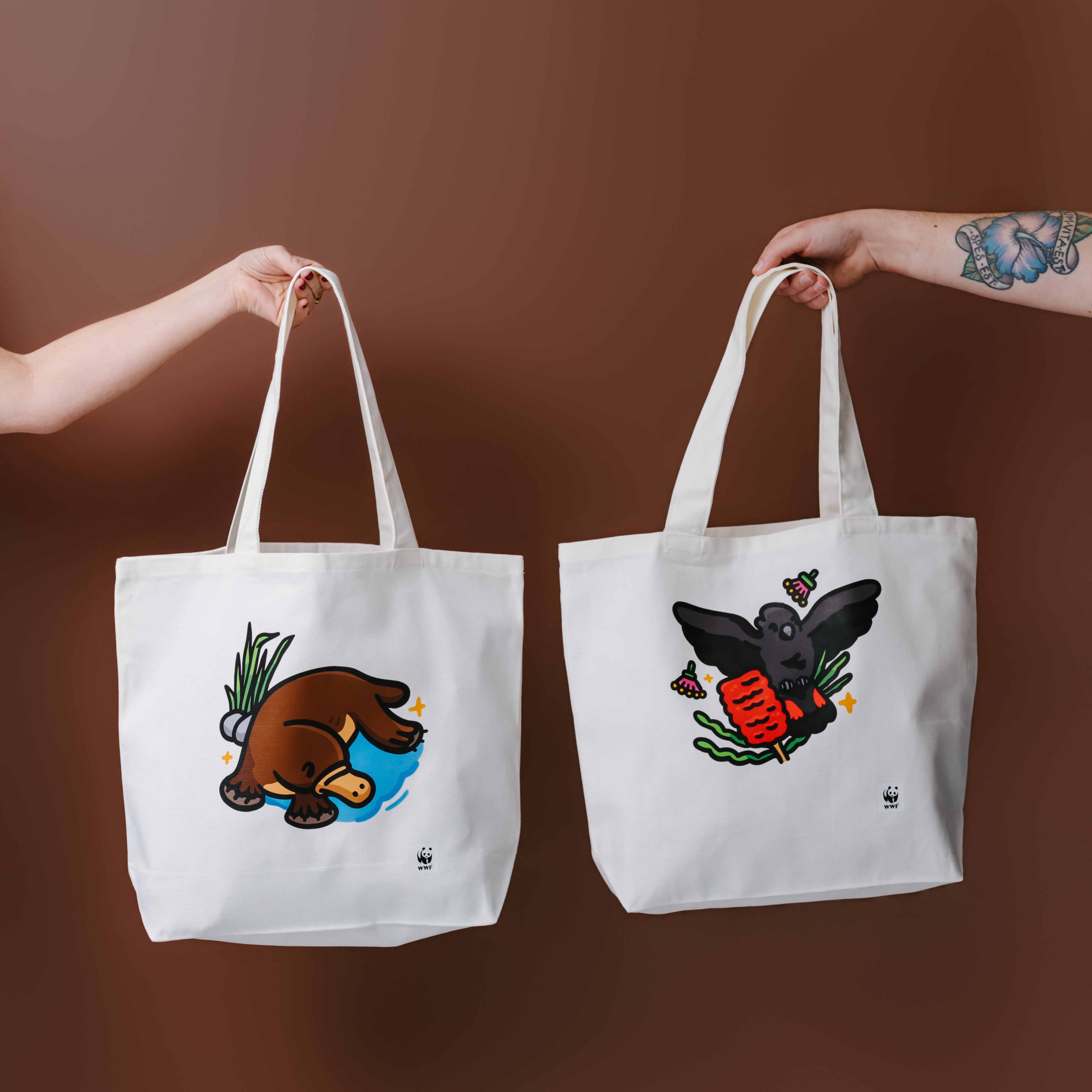 Two white canvas tote bags held up by human arms against a solid brown background. The tote bag on the left features a platypus illustration in a blue pond, and the tote bag on the right features a glossy black cockatoo illustration.