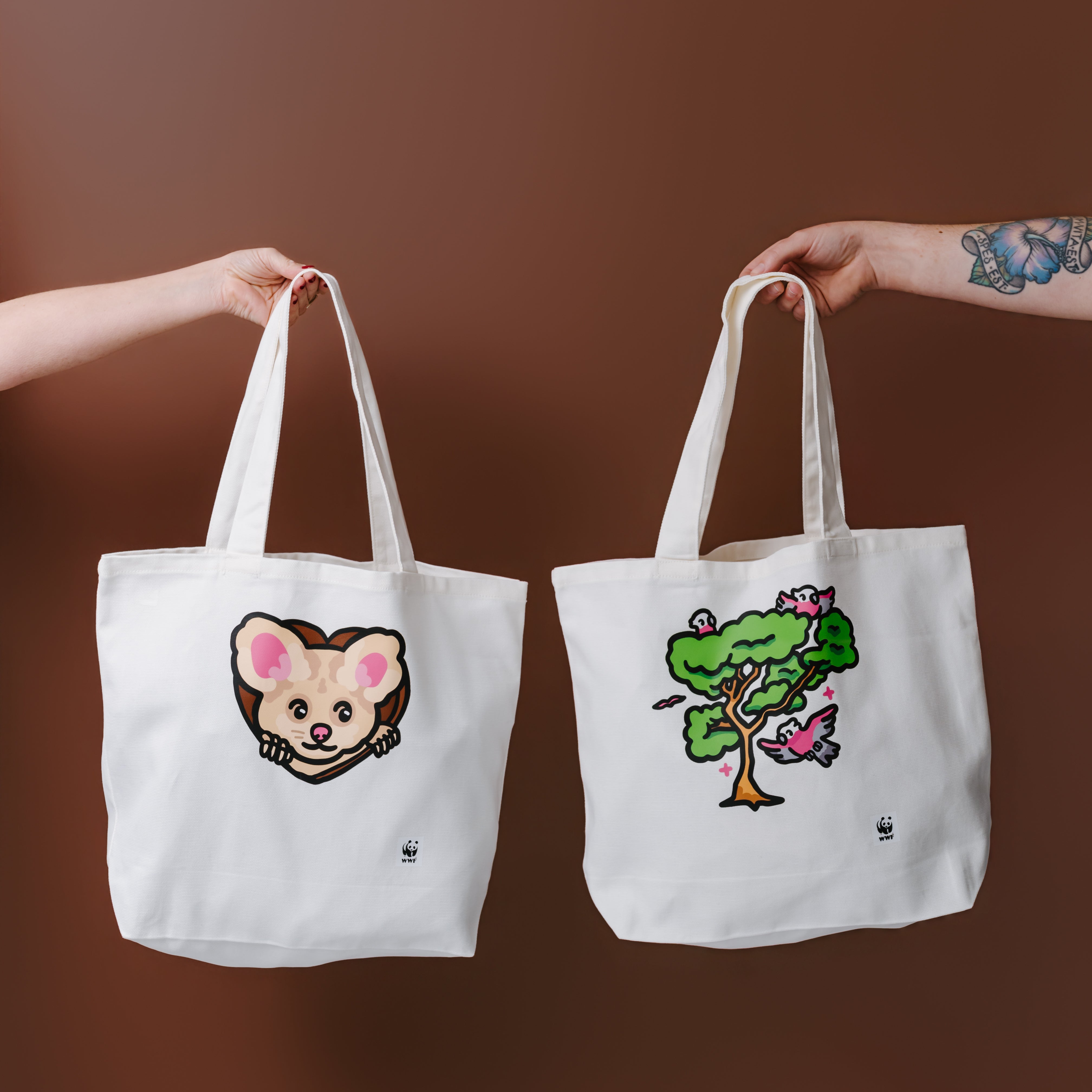 Two white canvas tote bags held up by human arms against a solid brown background. The tote bag on the left features a greater glider illustration, while the tote bag on the right features an illustrated tree with galas.