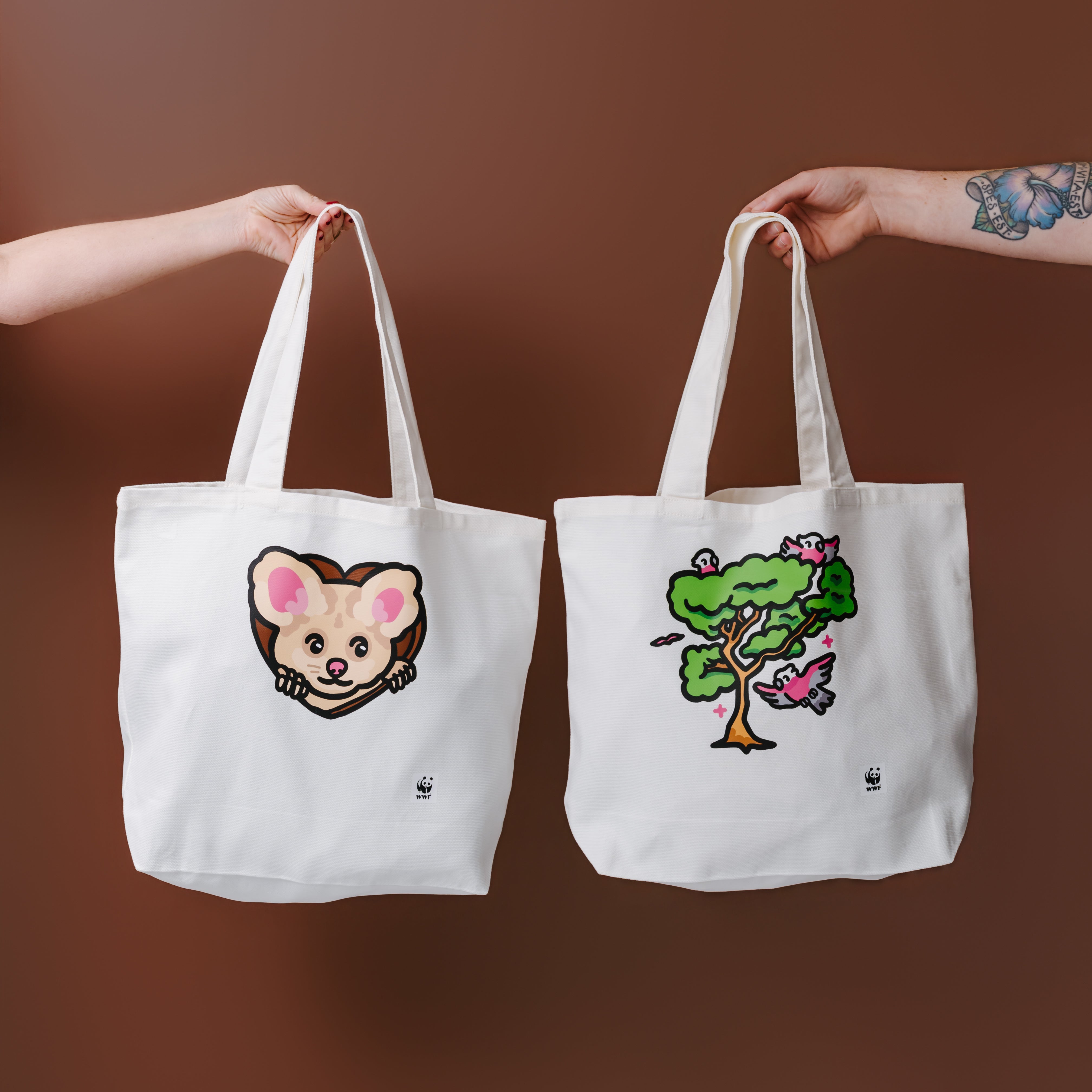 Two white canvas tote bags held up by human arms against a solid brown background. The left tote bag features a greater glider illustration, and the right tote bag features an illustrated tree with birds.