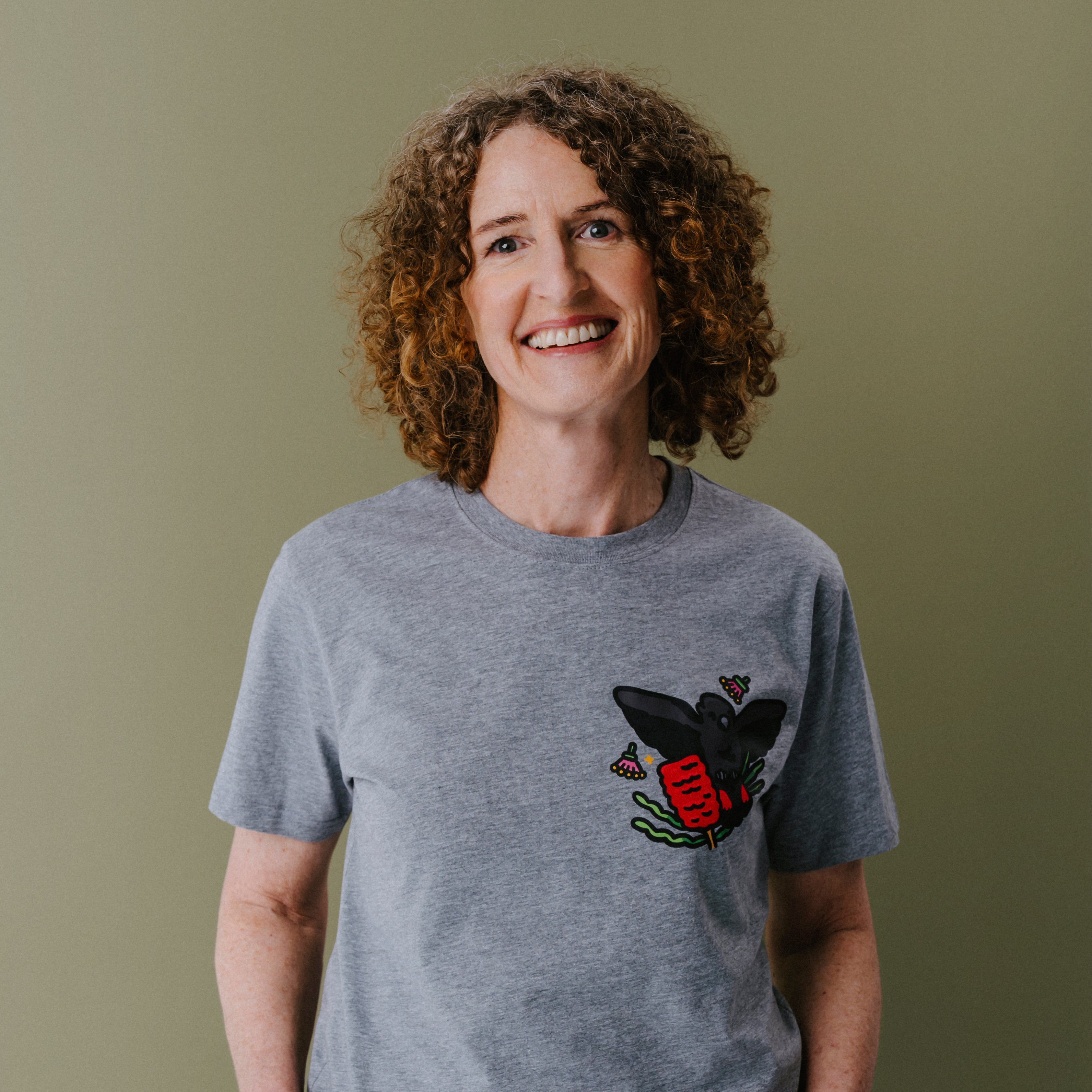 A smiling woman with curly brown hair wearing a heather gray t-shirt with a glossy black cockato illustration on the chest, set against a solid olive green background.