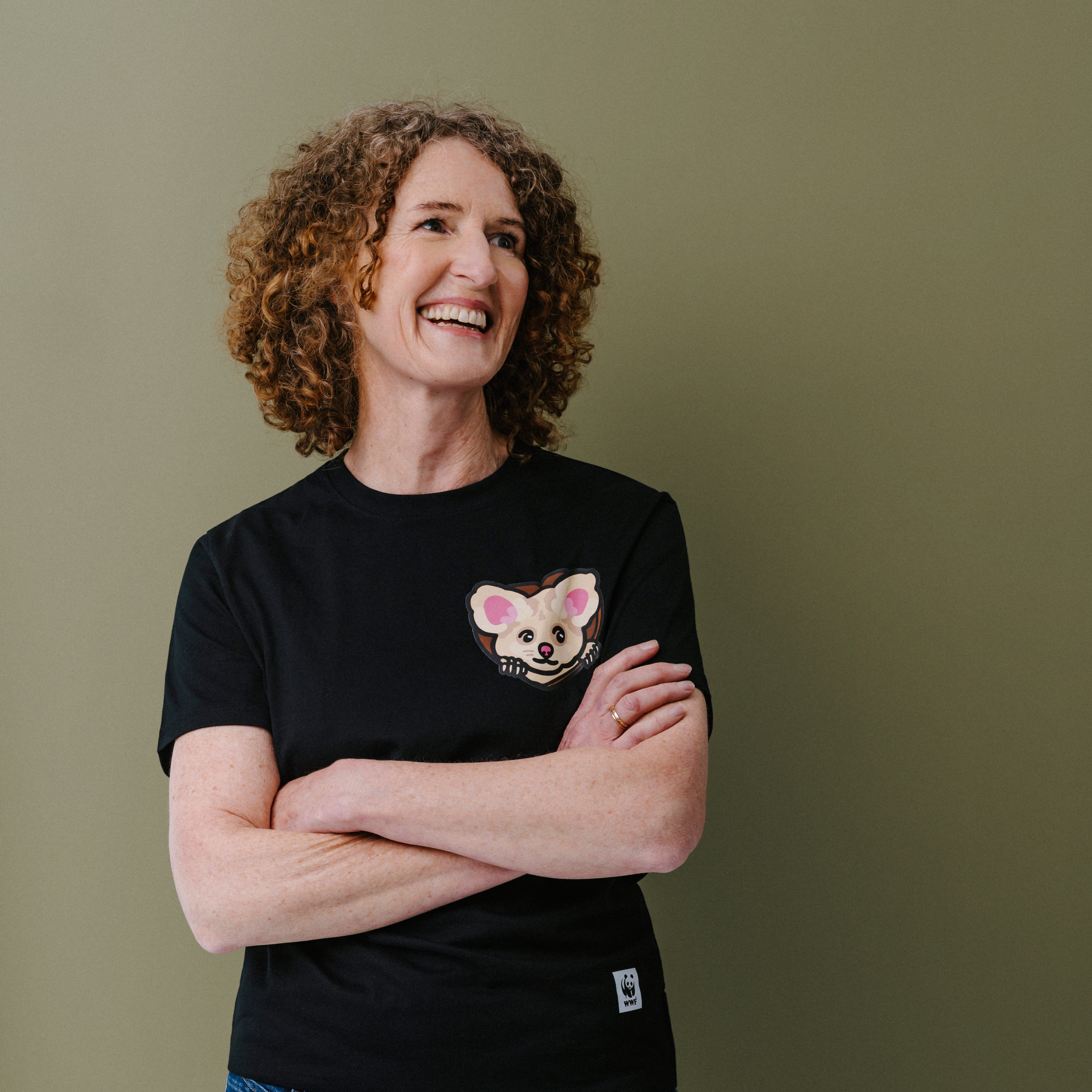 A smiling woman with curly brown hair stands with arms crossed, wearing a black short-sleeve T-shirt with a small illustrated greater glider face on the left chest and blue jeans, against a plain olive-green background.
