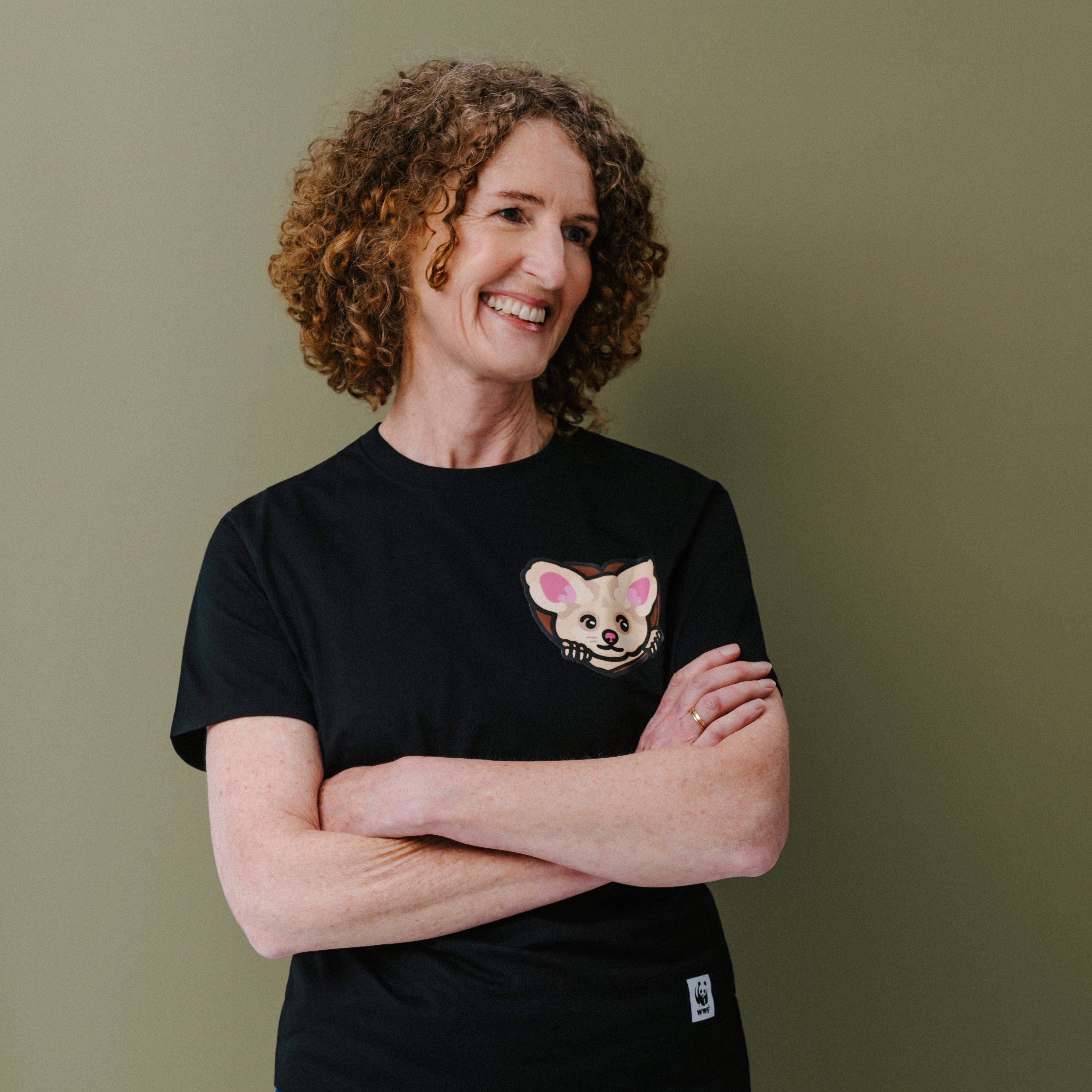 A smiling woman with curly brown hair stands with arms crossed, wearing a black short-sleeve T-shirt with a small illustrated greater glider face on the left chest and blue jeans, against a plain olive-green background.