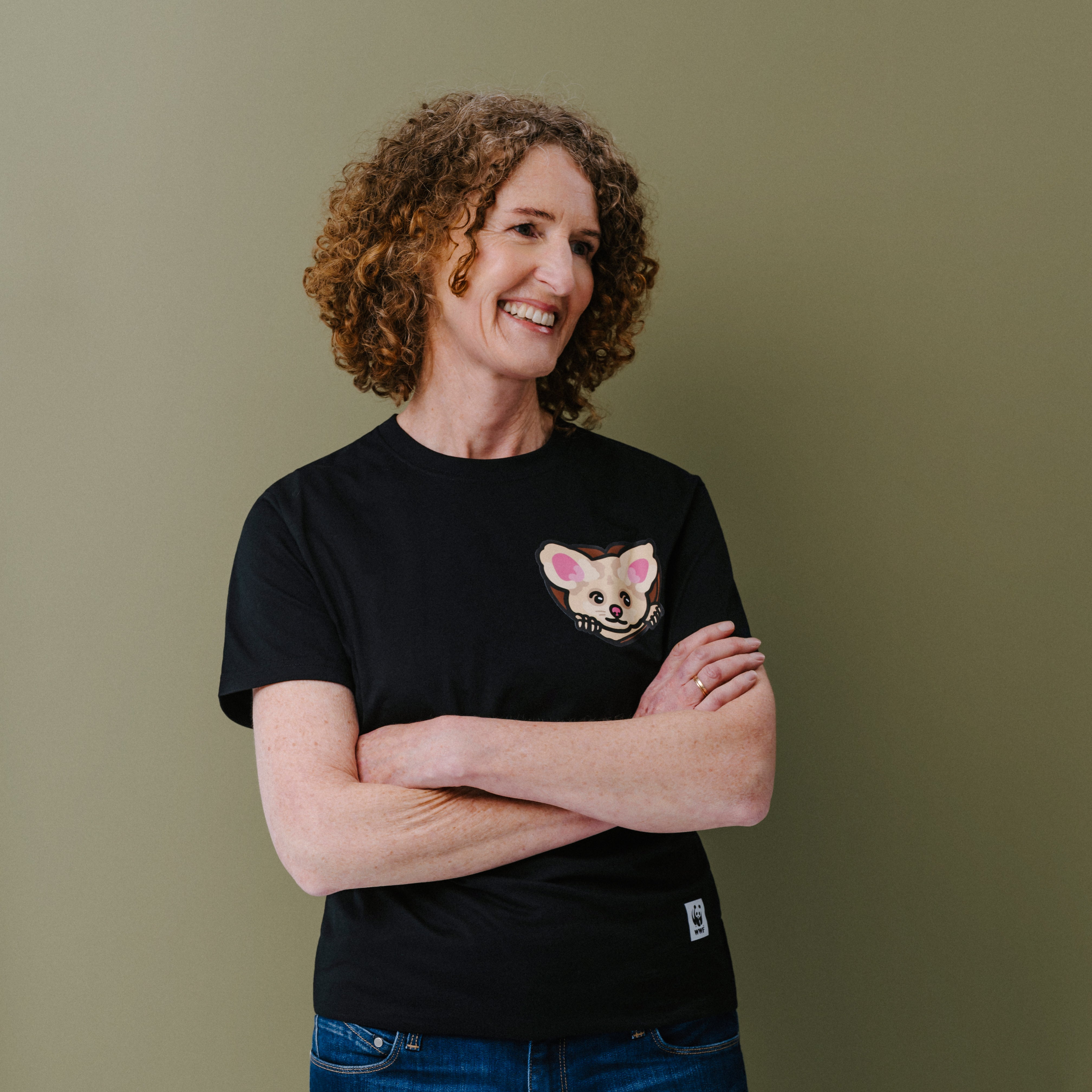 A smiling woman with curly brown hair stands with arms crossed, wearing a black short-sleeve T-shirt with a small illustrated greater glider face on the left chest and blue jeans, against a plain olive-green background.