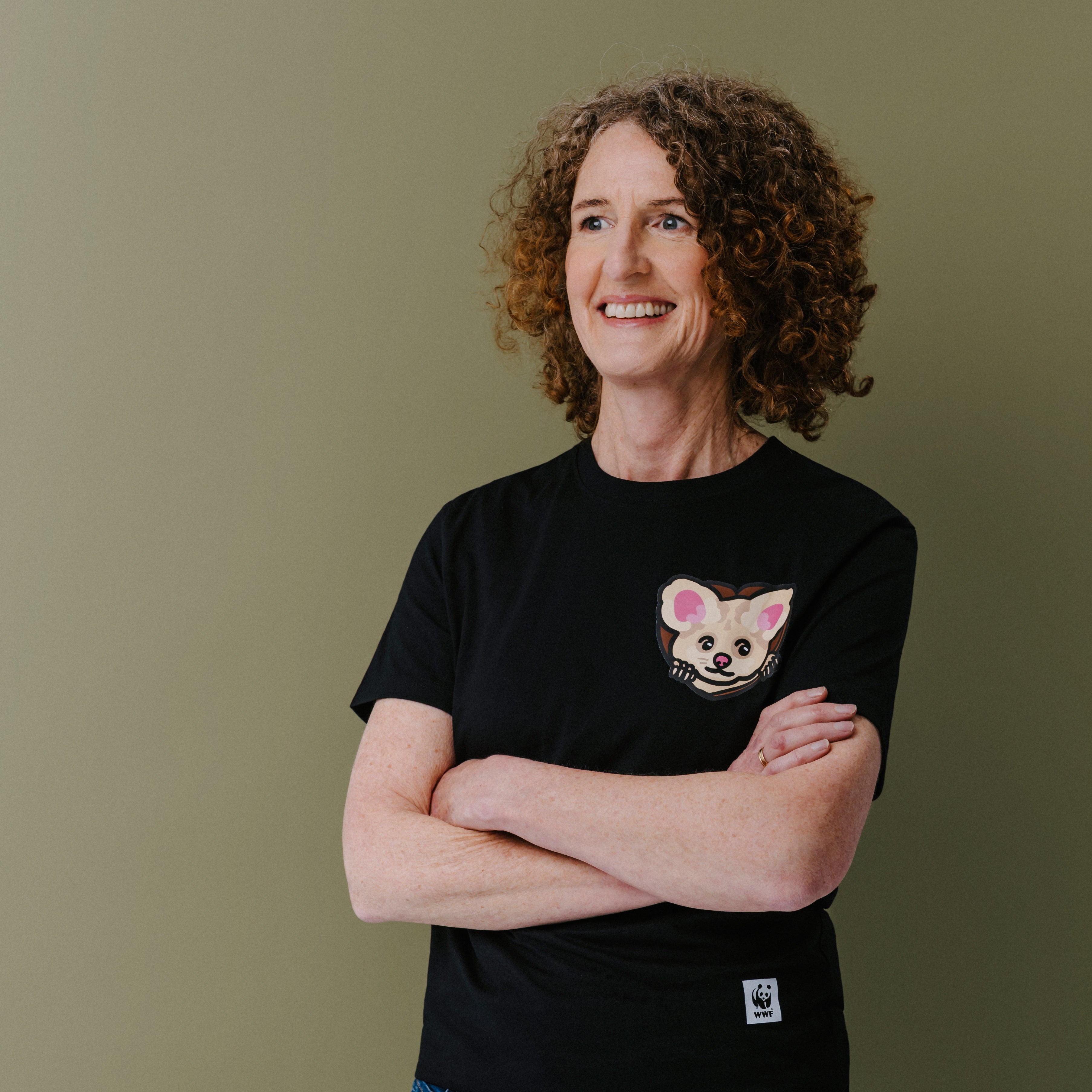 A smiling woman with curly brown hair stands with arms crossed, wearing a black short-sleeve T-shirt with a small illustrated greater glider face on the left chest and blue jeans, against a plain olive-green background.