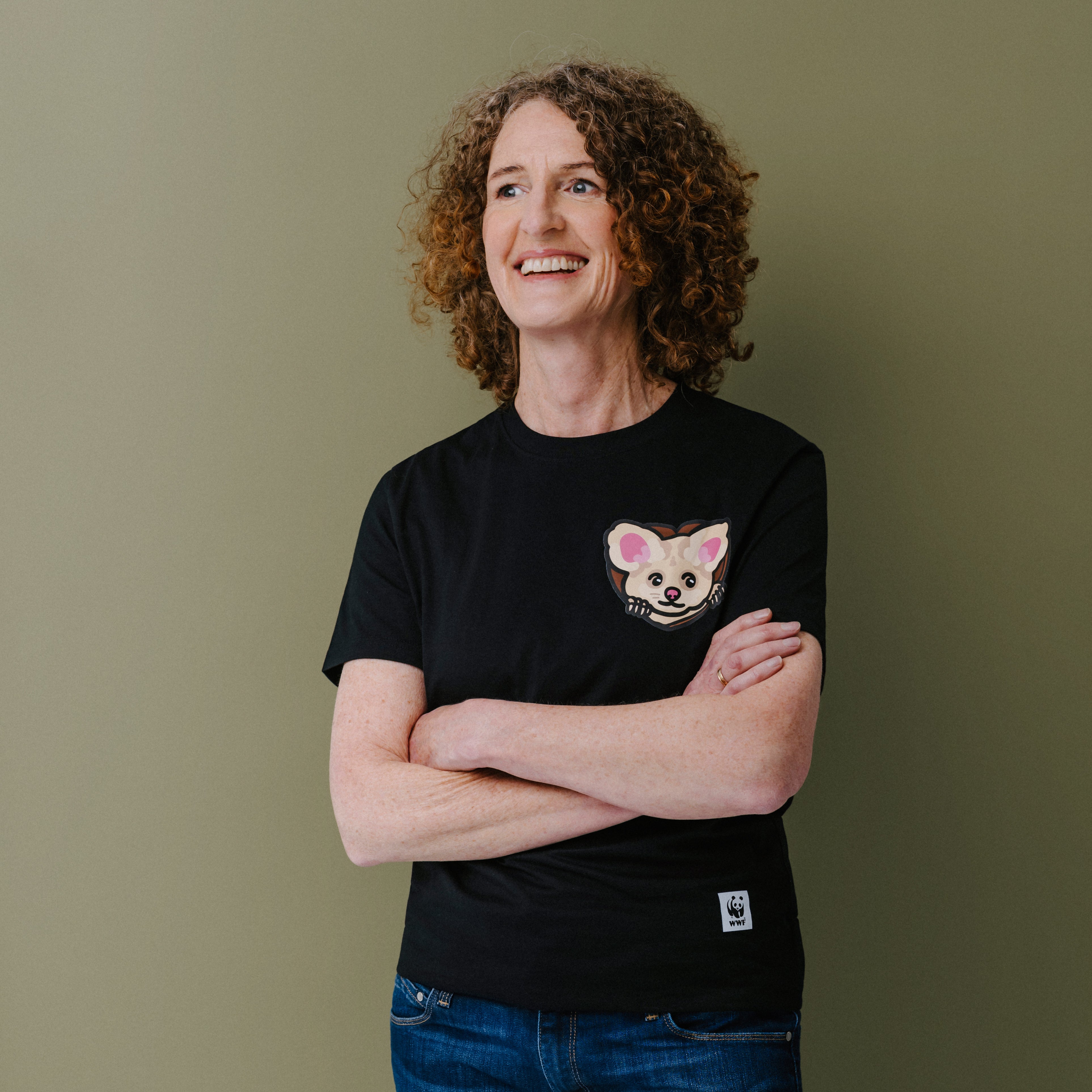 A smiling woman with curly brown hair stands with arms crossed, wearing a black short-sleeve T-shirt with a small illustrated greater glider face on the left chest and blue jeans, against a plain olive-green background.