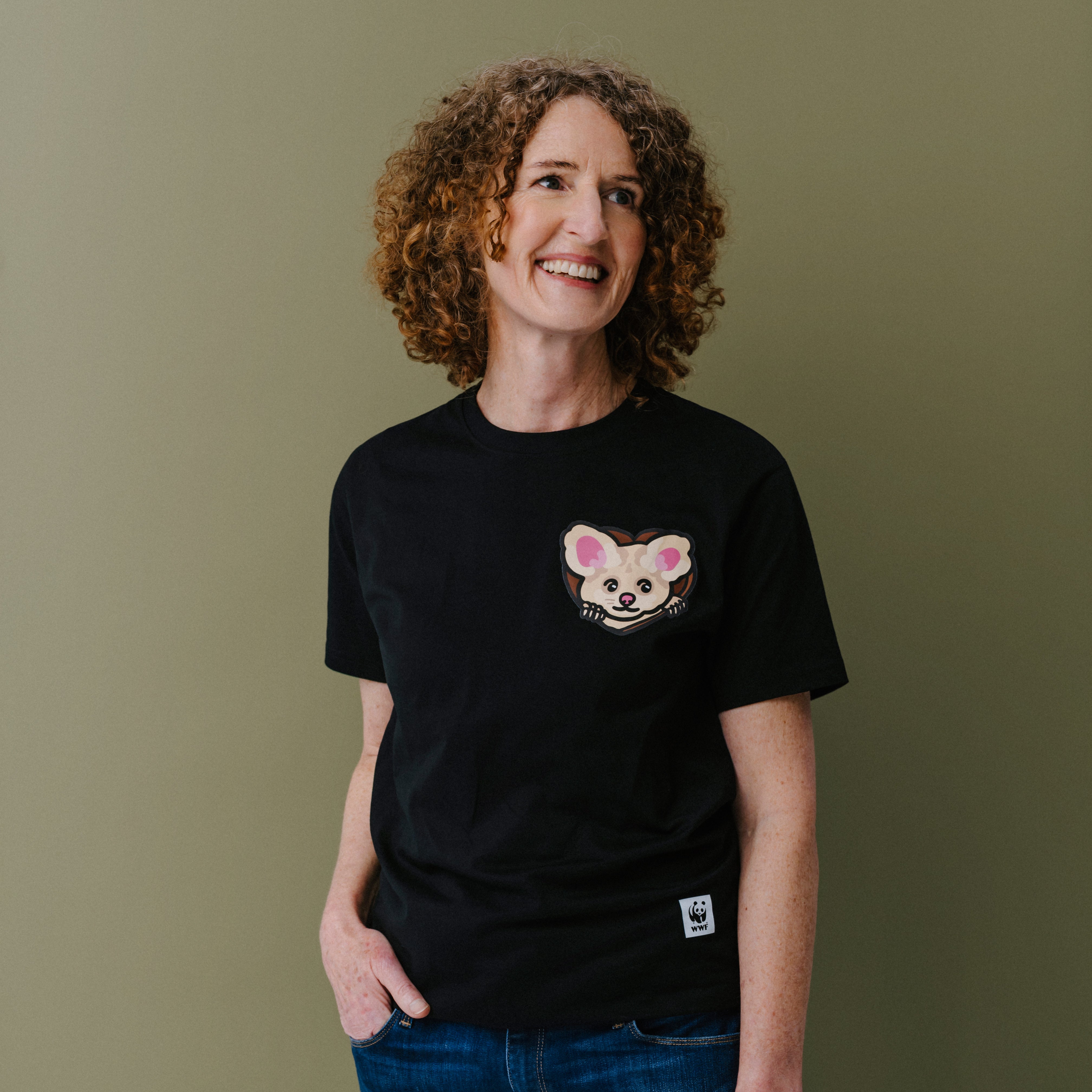 A smiling woman with curly brown hair stands with arms crossed, wearing a black short-sleeve T-shirt with a small illustrated greater glider face on the left chest and blue jeans, against a plain olive-green background.
