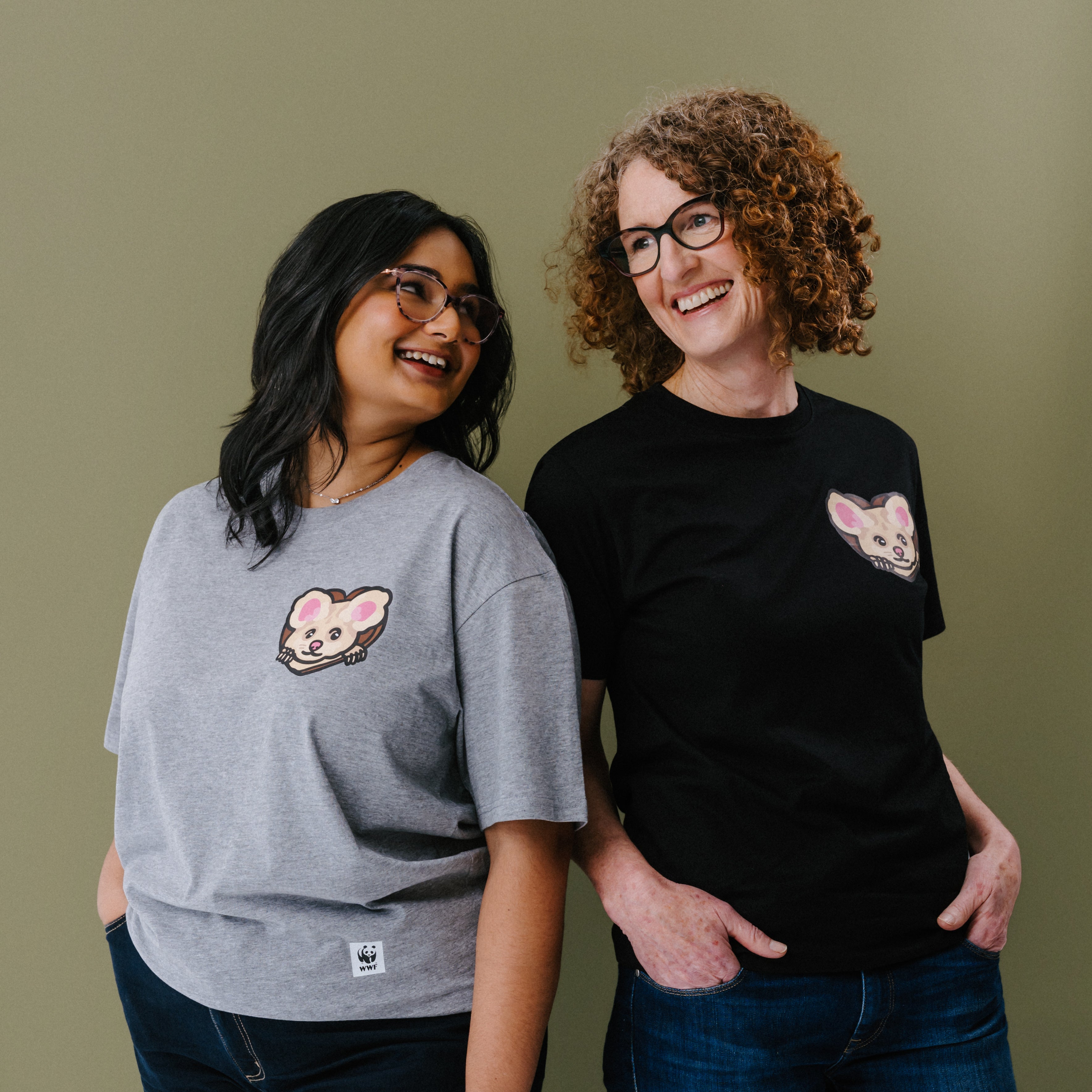 Two women standing side-by-side wearing matching graphic t-shirts with an illustrated greater glider on the left chest. The woman on the left wears a heather gray t-shirt, and the woman on the right wears a black t-shirt.