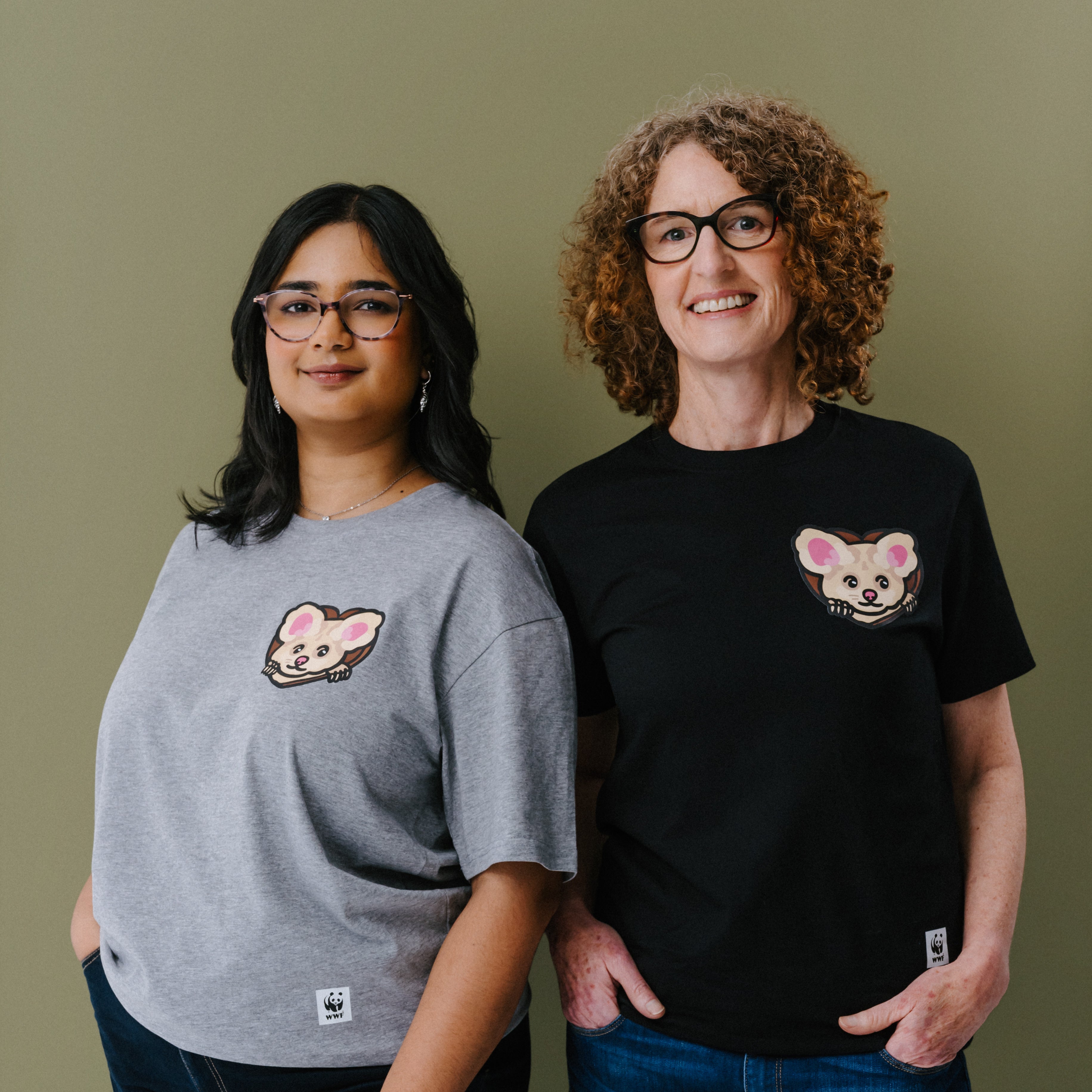 Two women standing side-by-side wearing matching graphic t-shirts with an illustrated greater glider on the left chest. The woman on the left wears a heather gray t-shirt, and the woman on the right wears a black t-shirt.