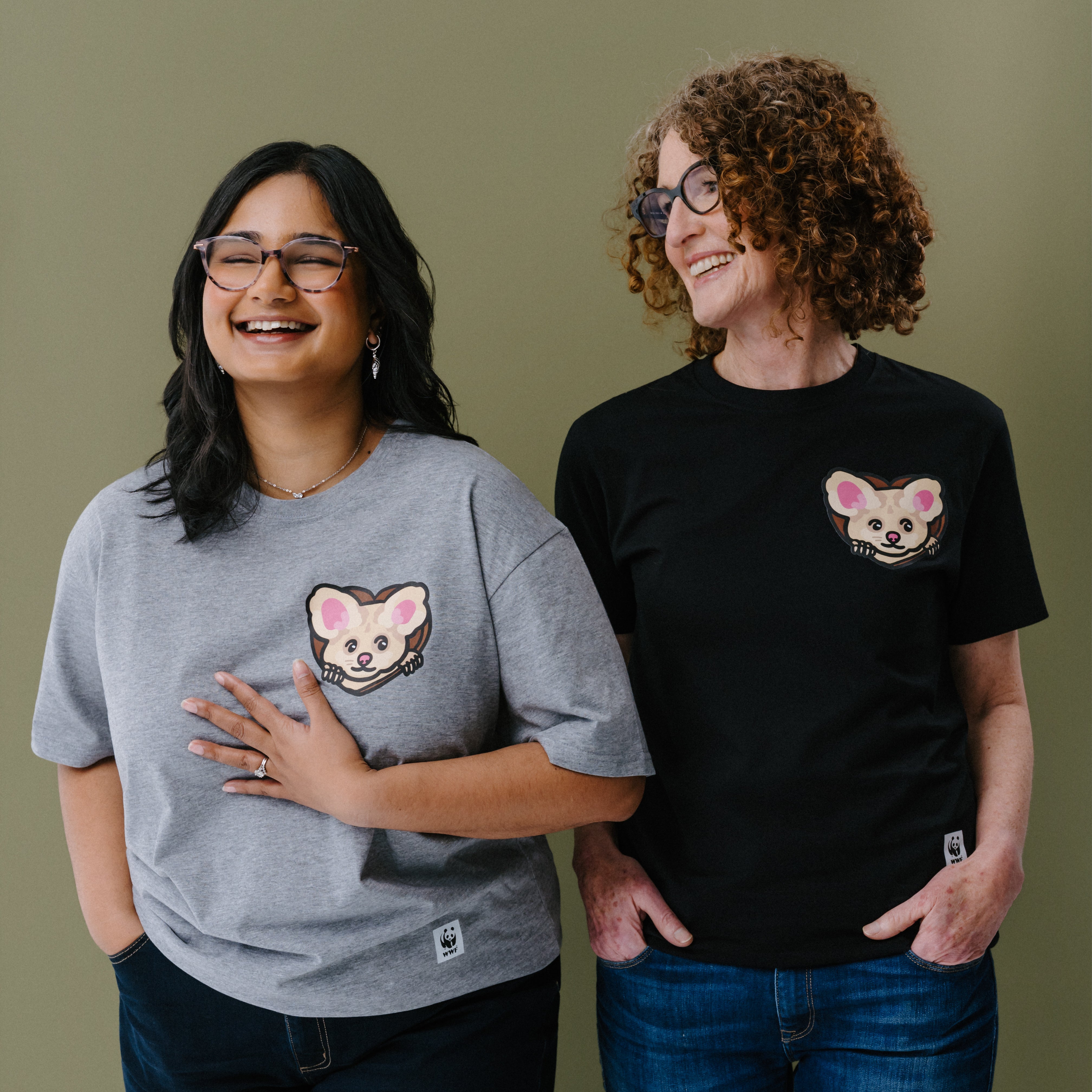 Two women standing side-by-side wearing matching graphic t-shirts with an illustrated greater glider on the left chest. The woman on the left wears a heather gray t-shirt, and the woman on the right wears a black t-shirt.