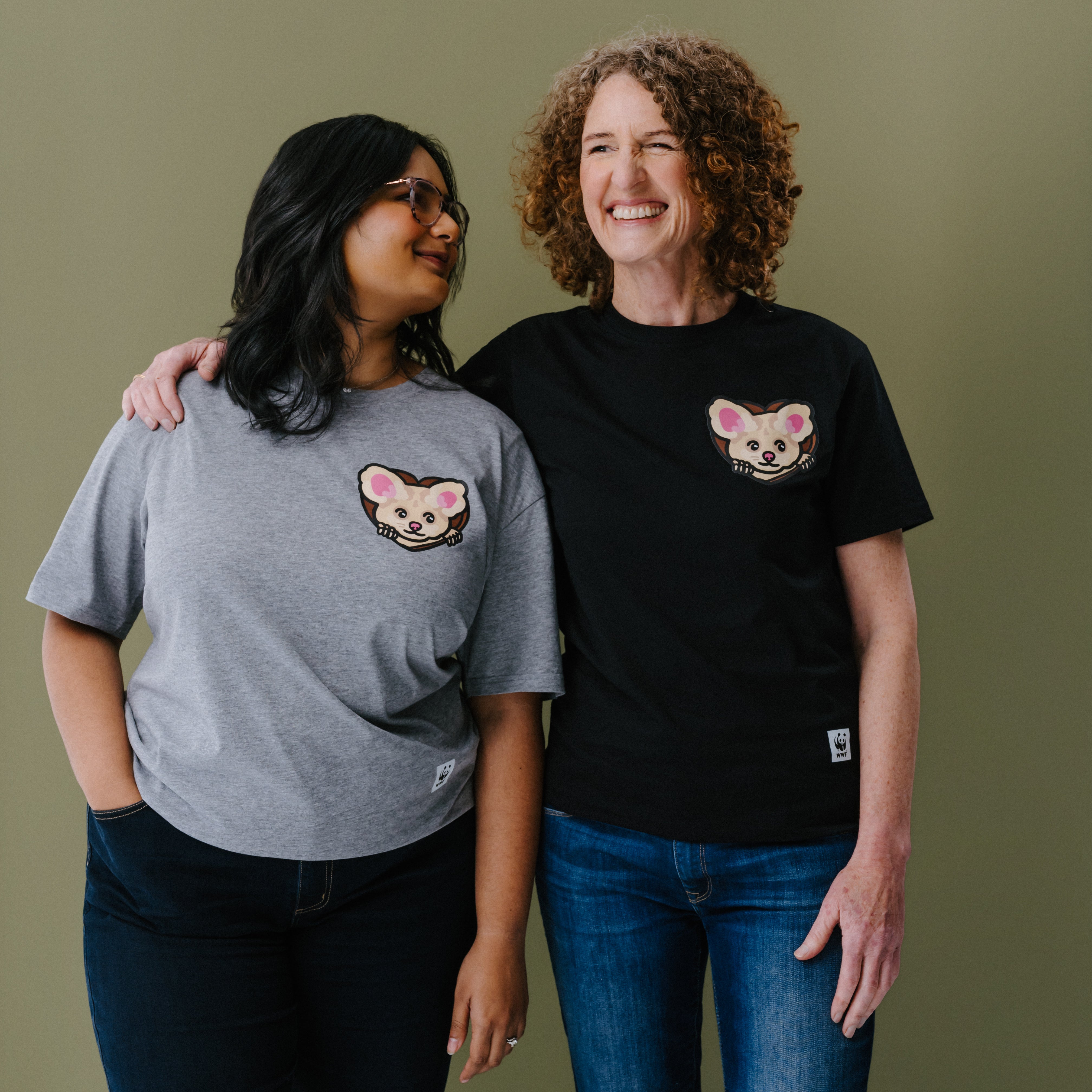Two women standing side-by-side wearing matching graphic t-shirts with an illustrated greater glider on the left chest. The woman on the left wears a heather gray t-shirt, and the woman on the right wears a black t-shirt.