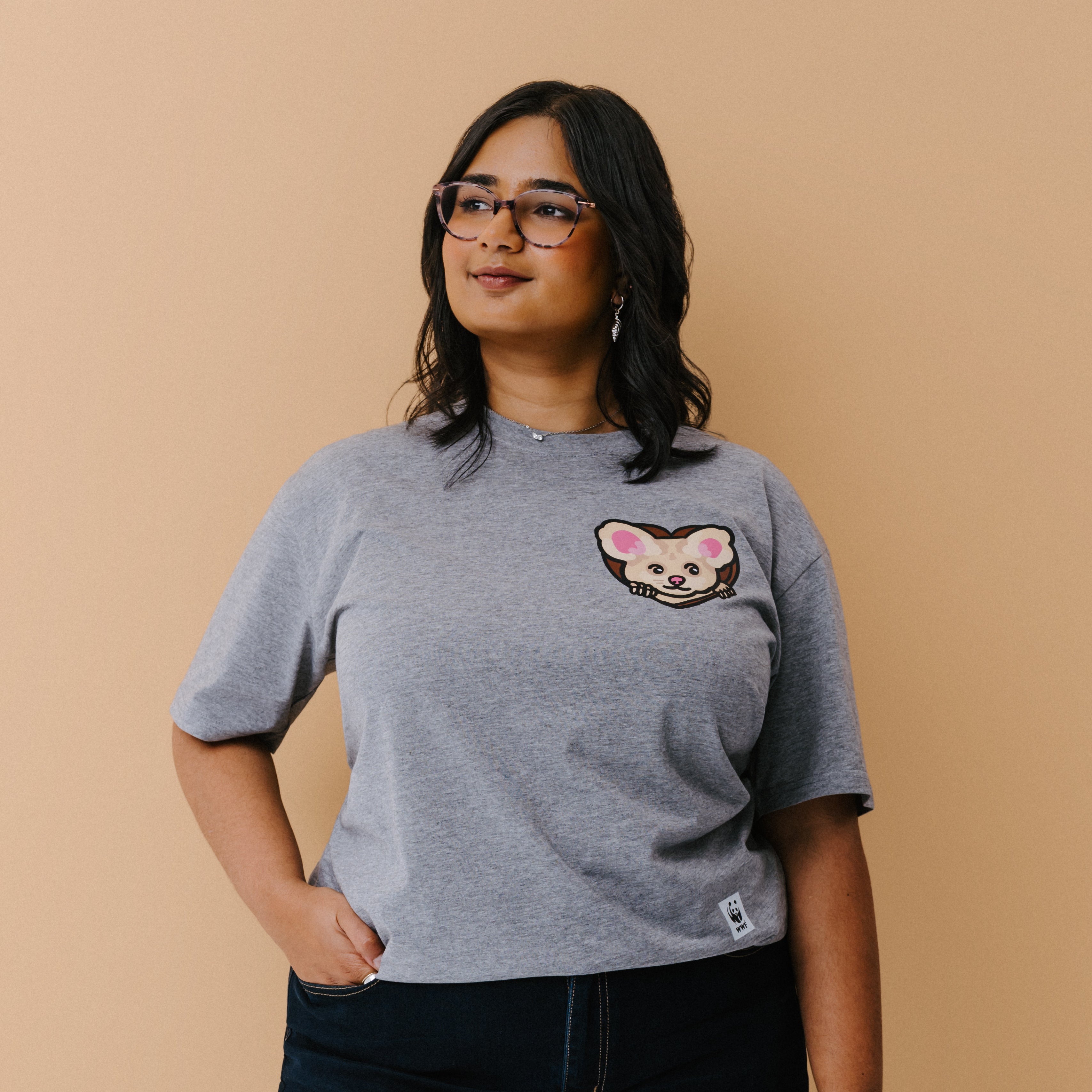 A woman with glasses is wearing a heather gray t-shirt with a small illustrated greater glider graphic on the chest, paired with dark pants, set against a solid beige background.