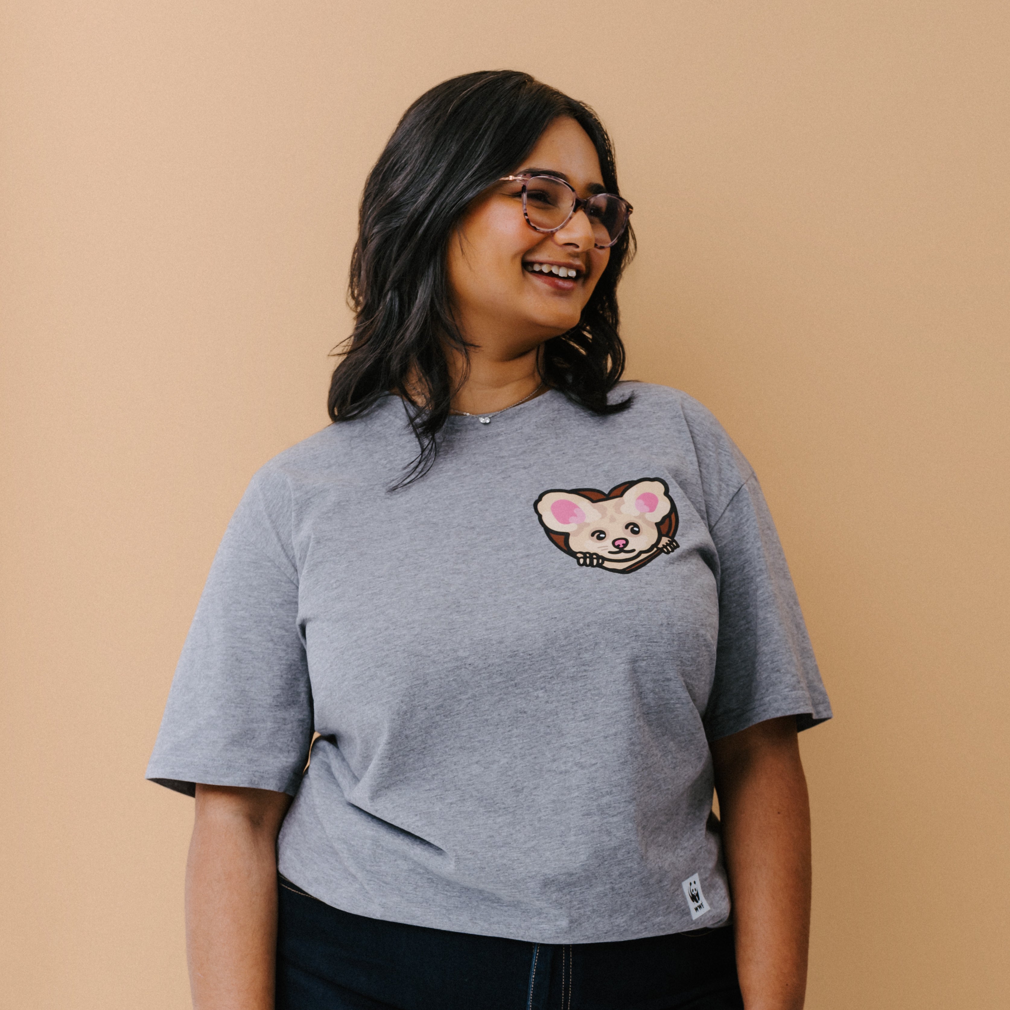 A woman with glasses is wearing a heather gray t-shirt with a small illustrated greater glider graphic on the chest, paired with dark pants, set against a solid beige background.