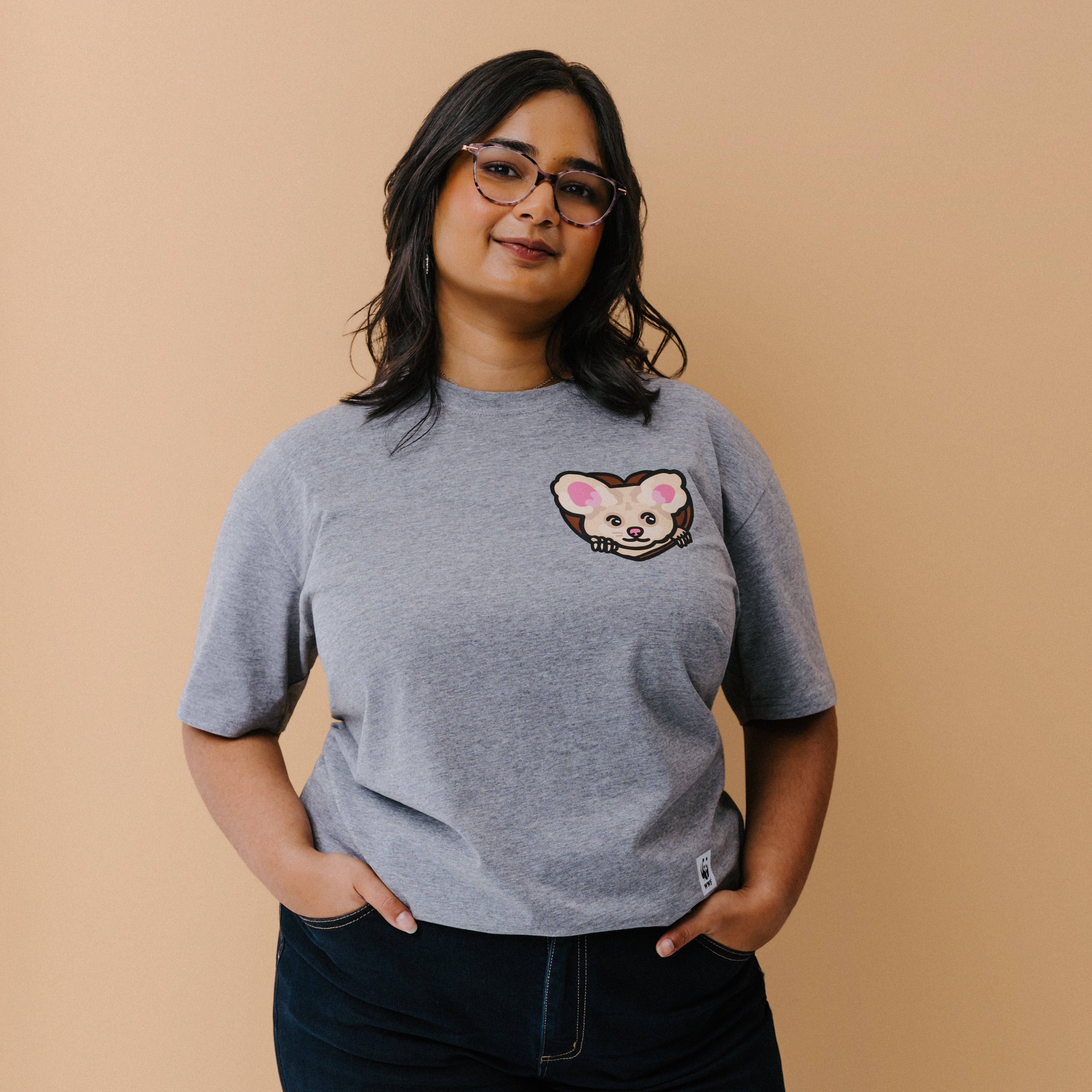 A woman with glasses is wearing a heather gray t-shirt with a small illustrated greater glider graphic on the chest, paired with dark pants, set against a solid beige background.