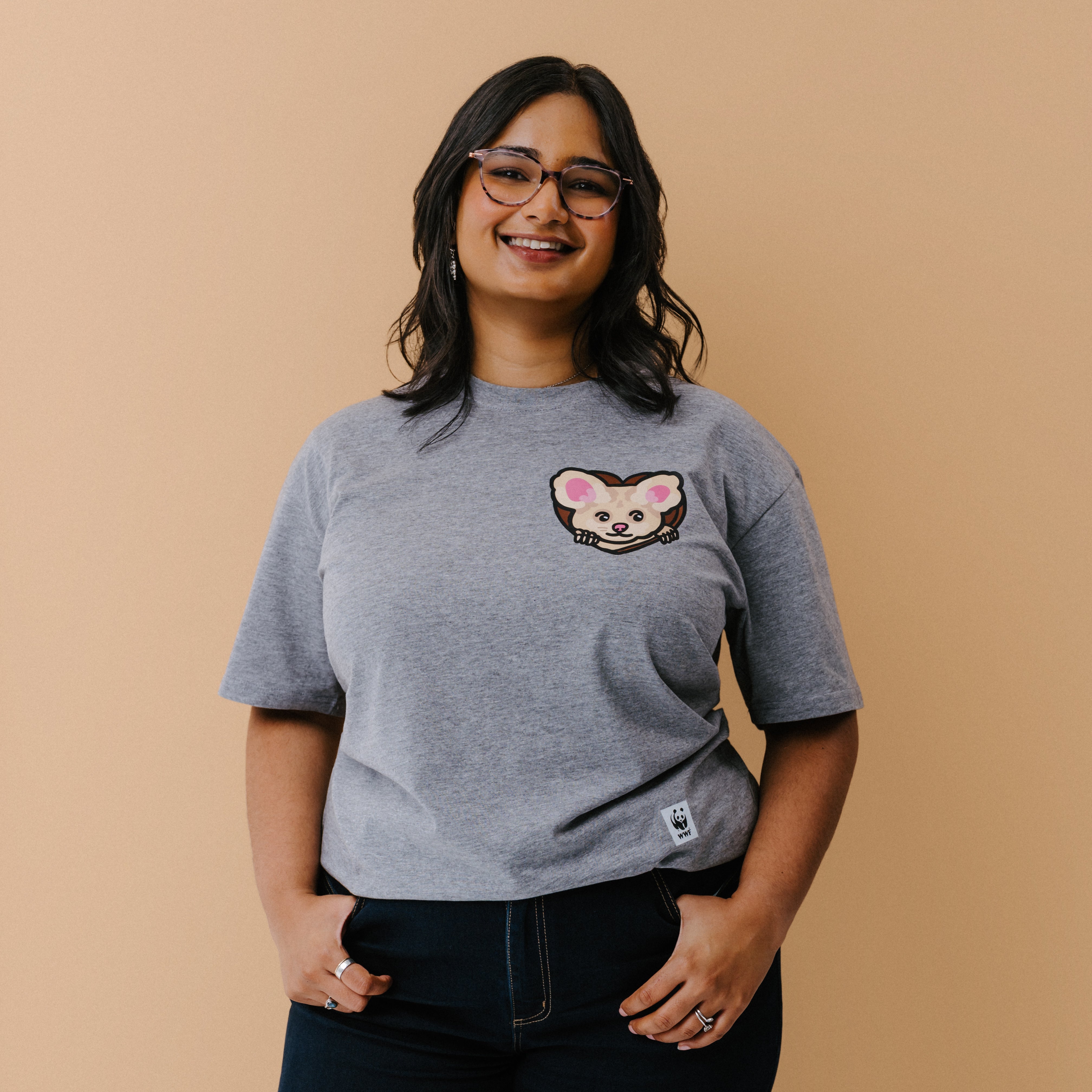 A woman with glasses is wearing a heather gray t-shirt with a small illustrated greater glider graphic on the chest, paired with dark pants, set against a solid beige background.