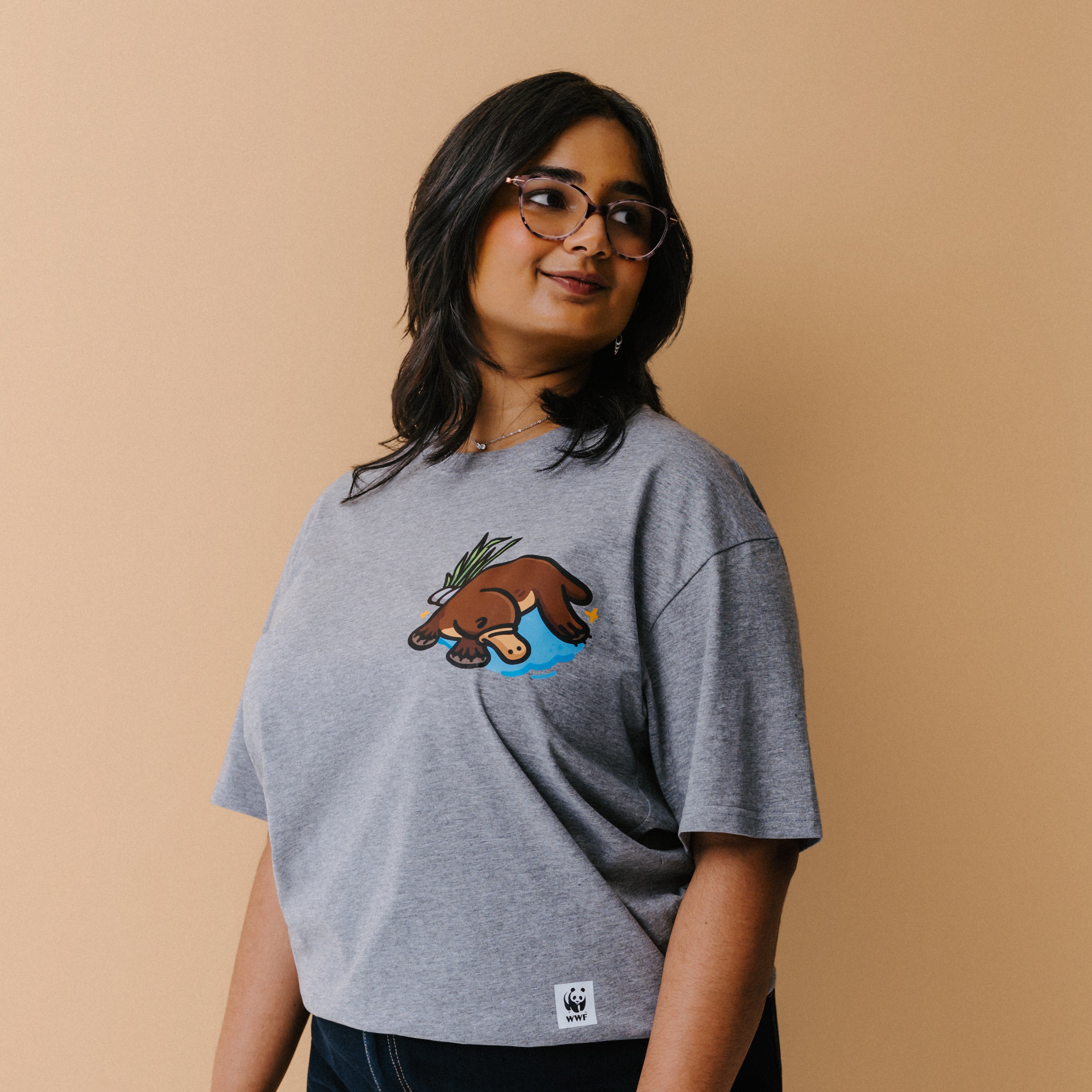 A woman with glasses is wearing a heather gray t-shirt with a small illustrated platypus in a blue pond and green plant on the chest, set against a solid beige background.