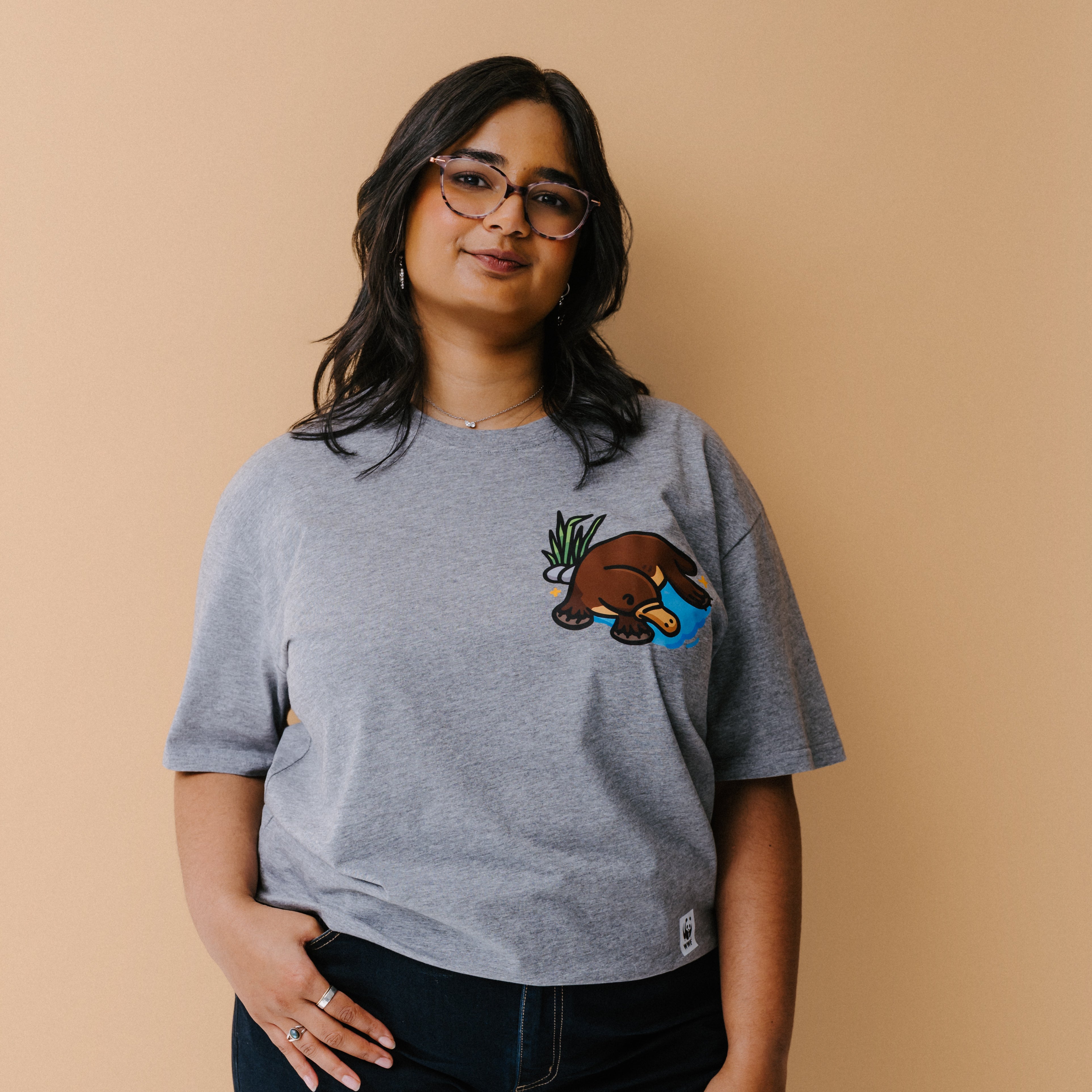 A woman with glasses is wearing a heather gray t-shirt with a small illustrated platypus in a blue pond and green plant on the chest, set against a solid beige background.