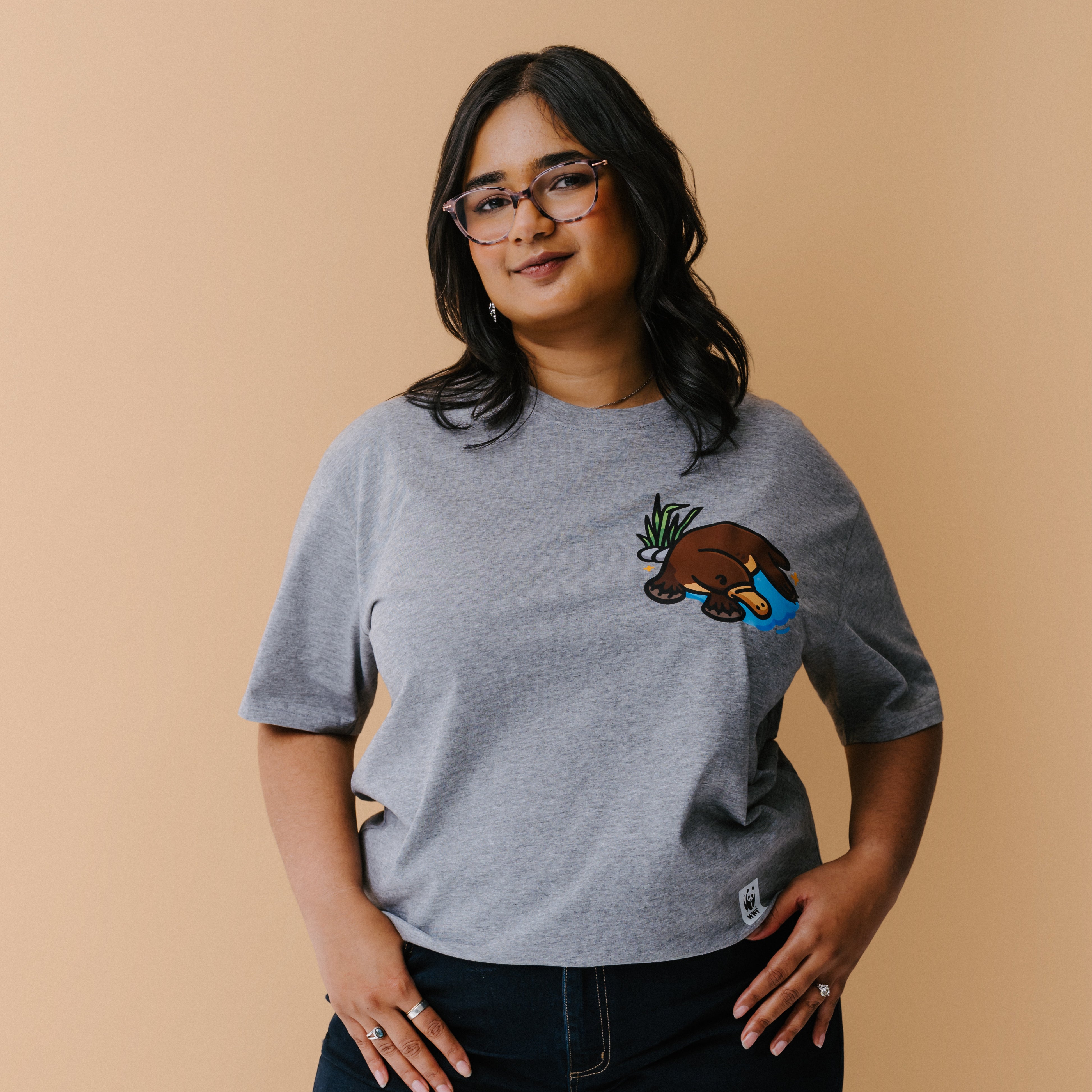 A woman with glasses is wearing a heather gray t-shirt with a small illustrated platypus in a blue pond and green plant on the chest, set against a solid beige background.