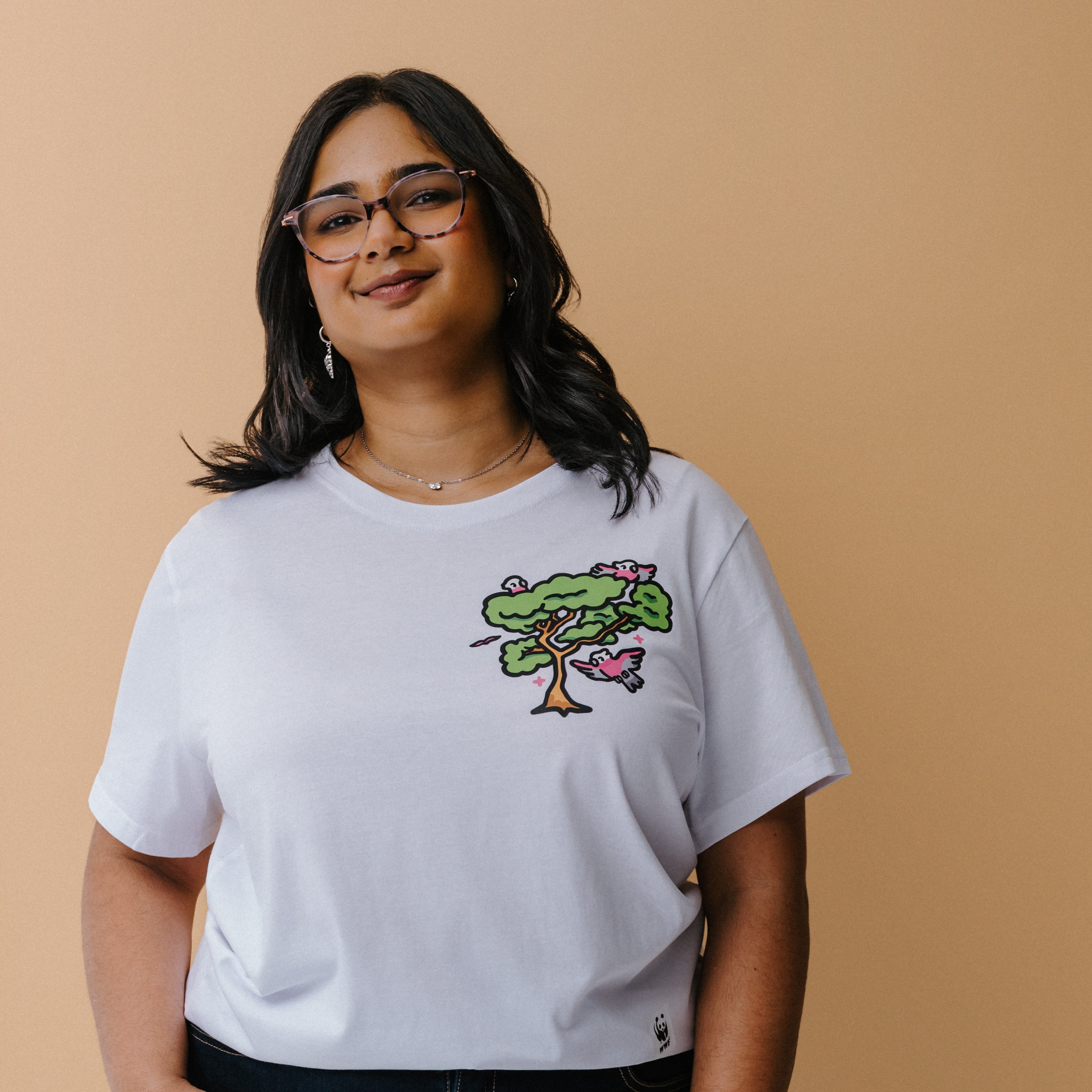 A woman with glasses and a necklace wearing a white t-shirt with a colorful tree and galas illustration on the chest, posed against a solid beige background.