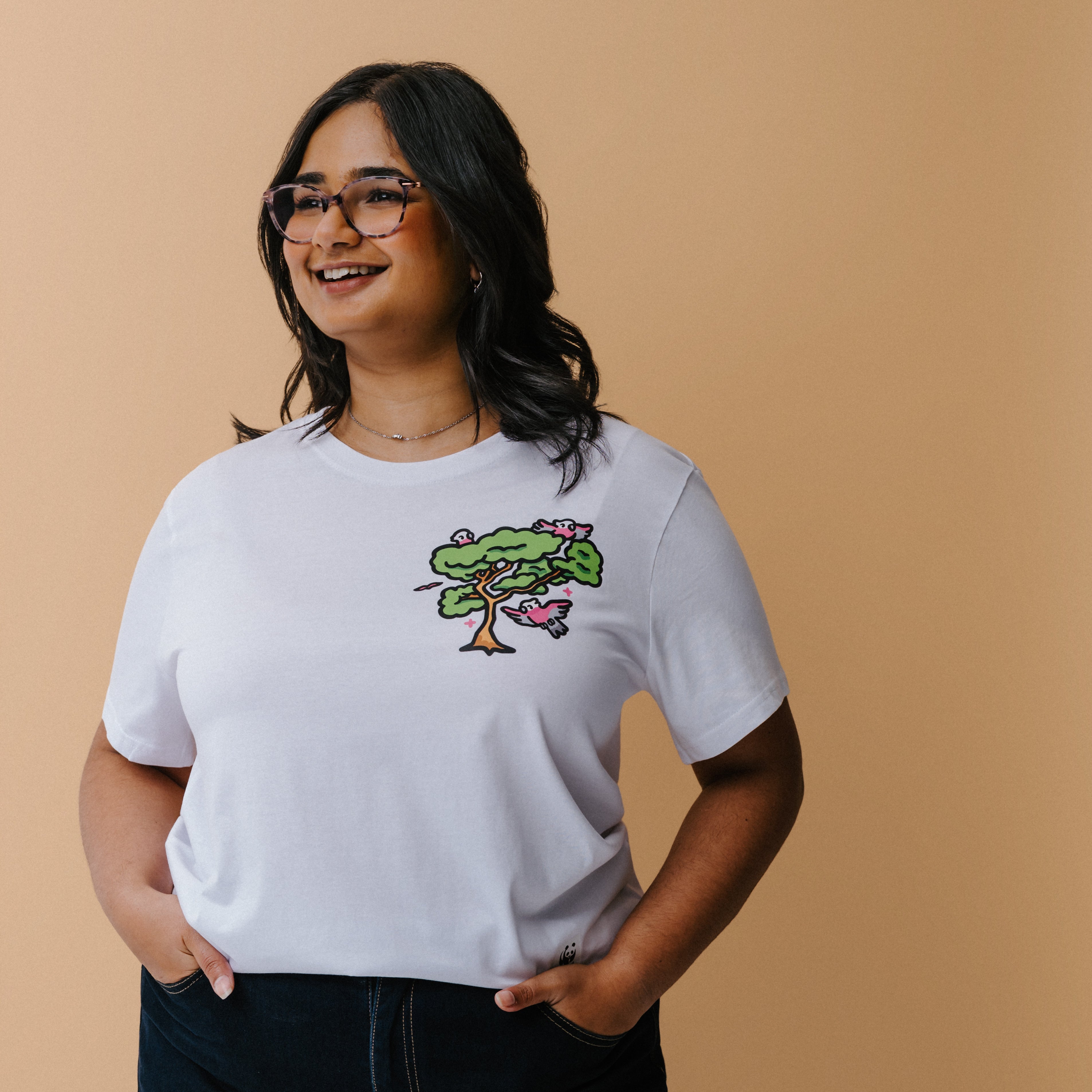 A woman with glasses and a necklace wearing a white t-shirt with a colorful tree and galas illustration on the chest, posed against a solid beige background.
