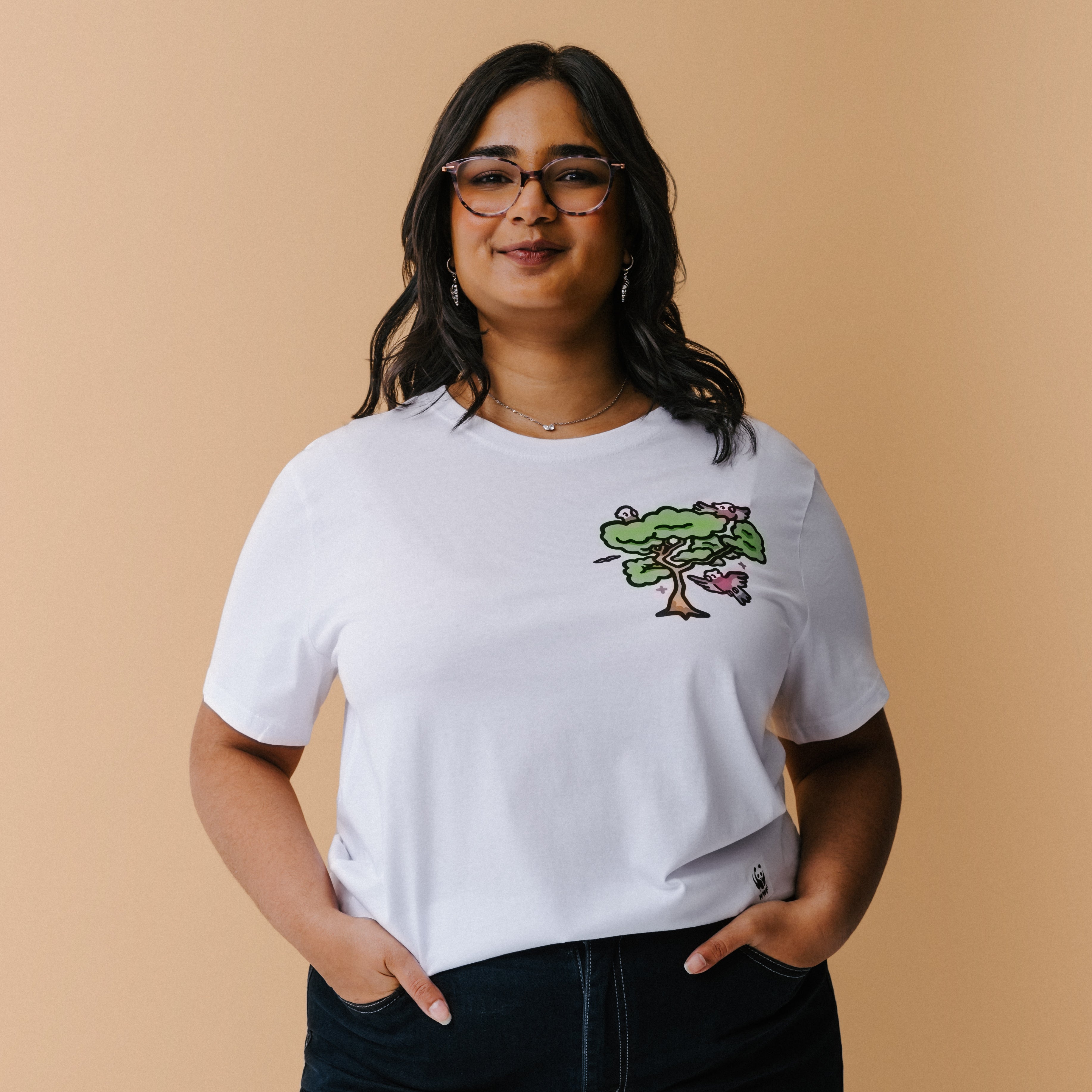 A woman with glasses and a necklace wearing a white t-shirt with a colorful tree and galas illustration on the chest, posed against a solid beige background.
