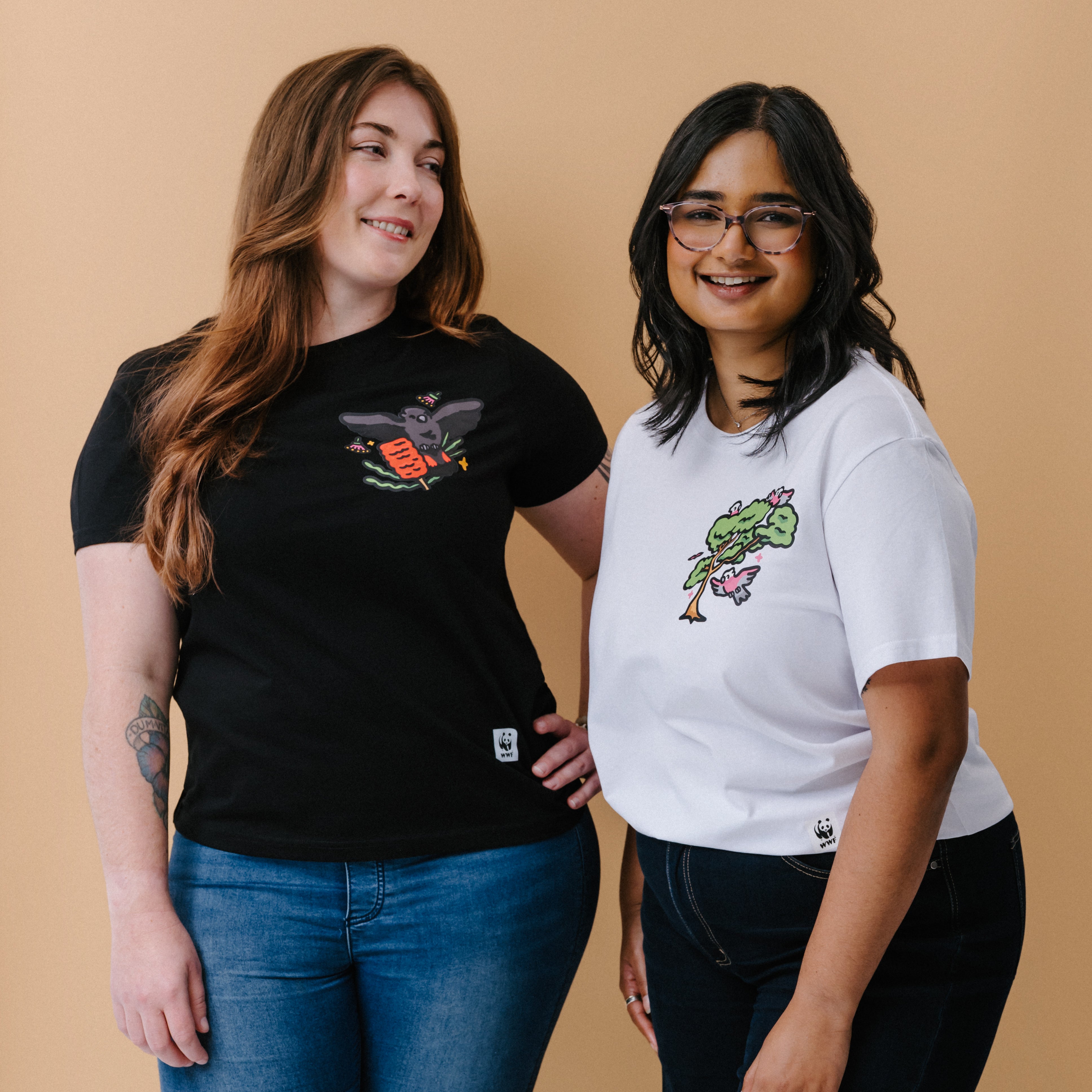 Two women standing side-by-side wearing graphic t-shirts. The woman on the left wears a black crewneck t-shirt with a glossy black cockatoo graphic and blue jeans.