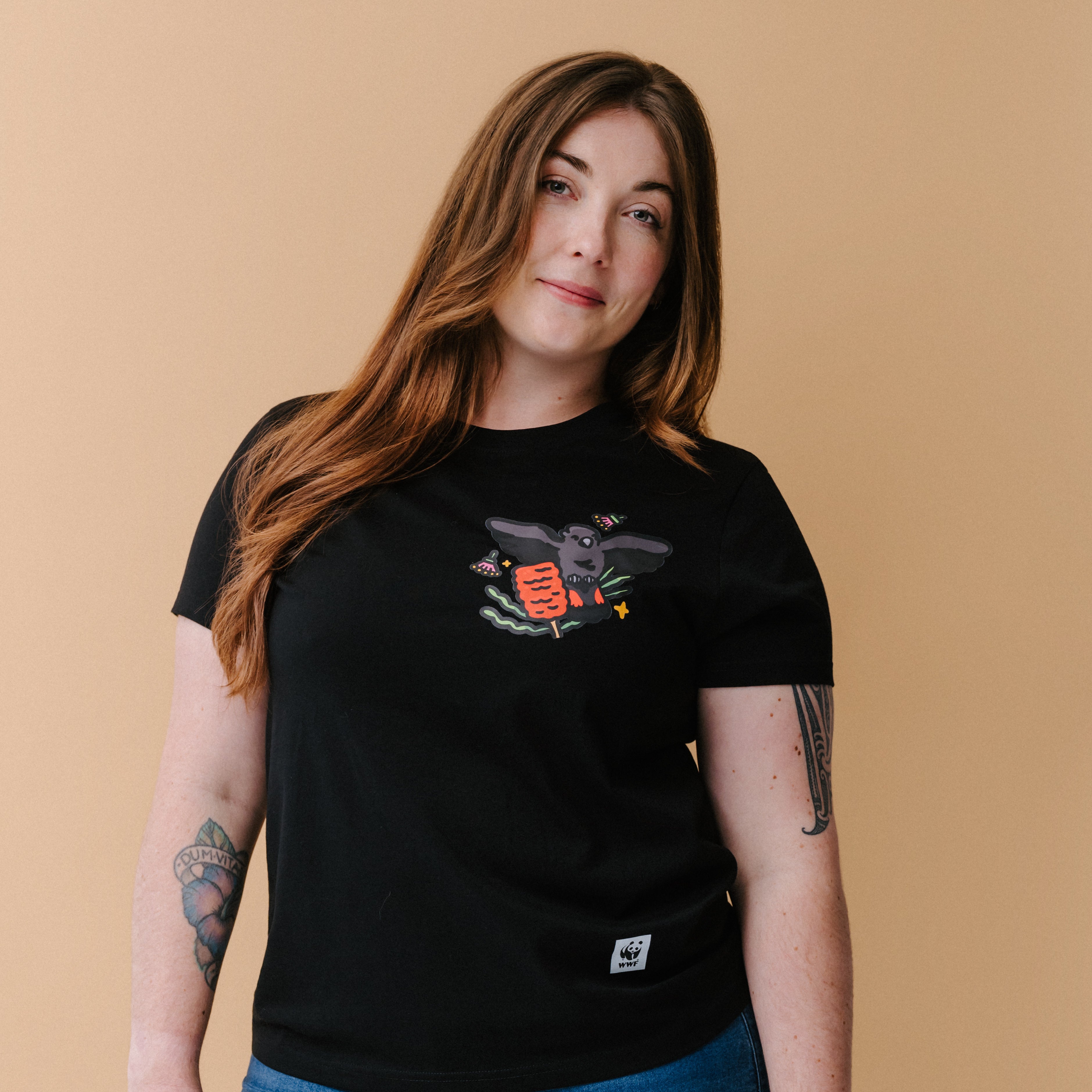 A woman with long brown hair and tattoos wearing a black graphic t-shirt with an illustrated glossy black cockatoo, posing against a solid beige background.