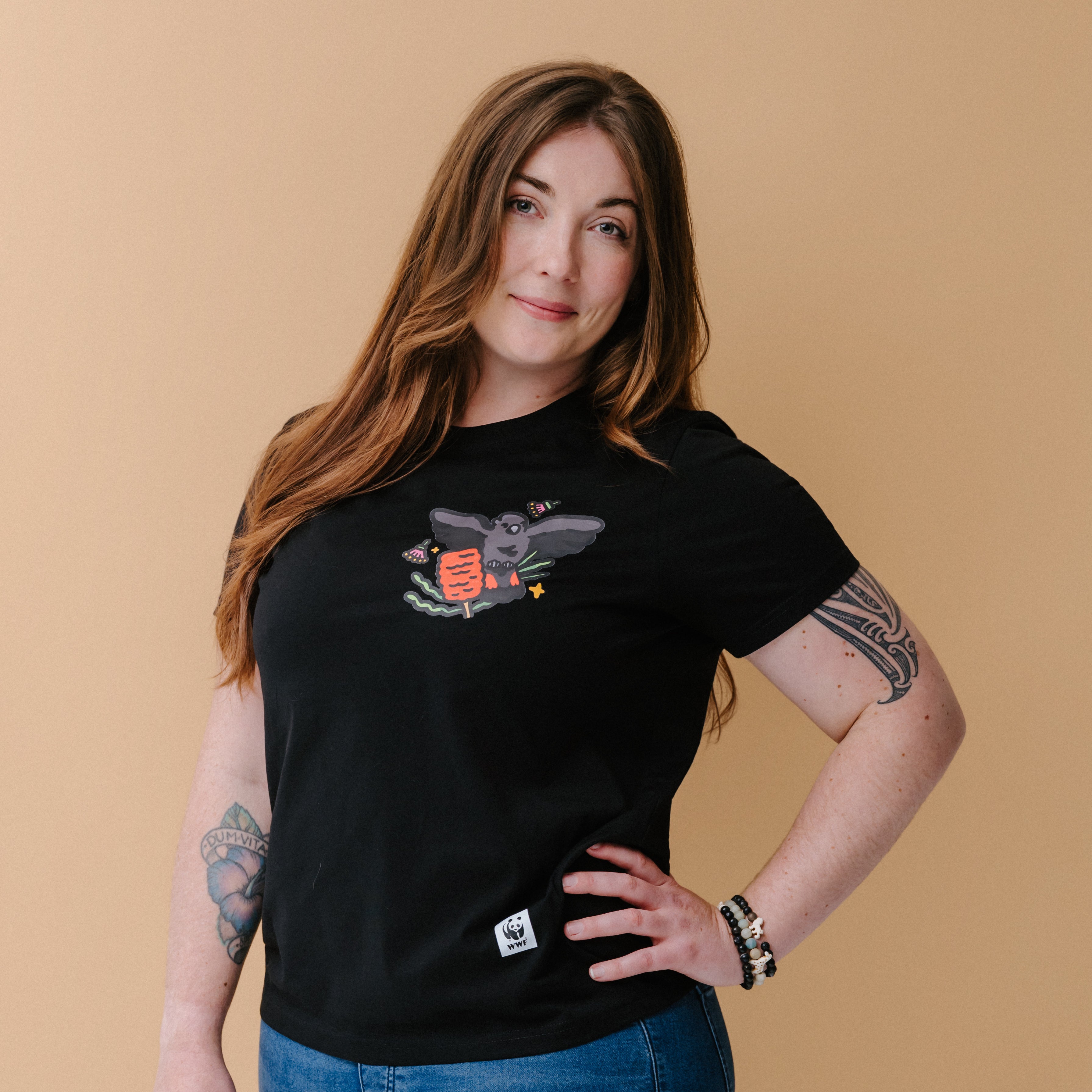 A woman with long brown hair and tattoos wearing a black graphic t-shirt with an illustrated glossy black cockatoo, posing against a solid beige background.