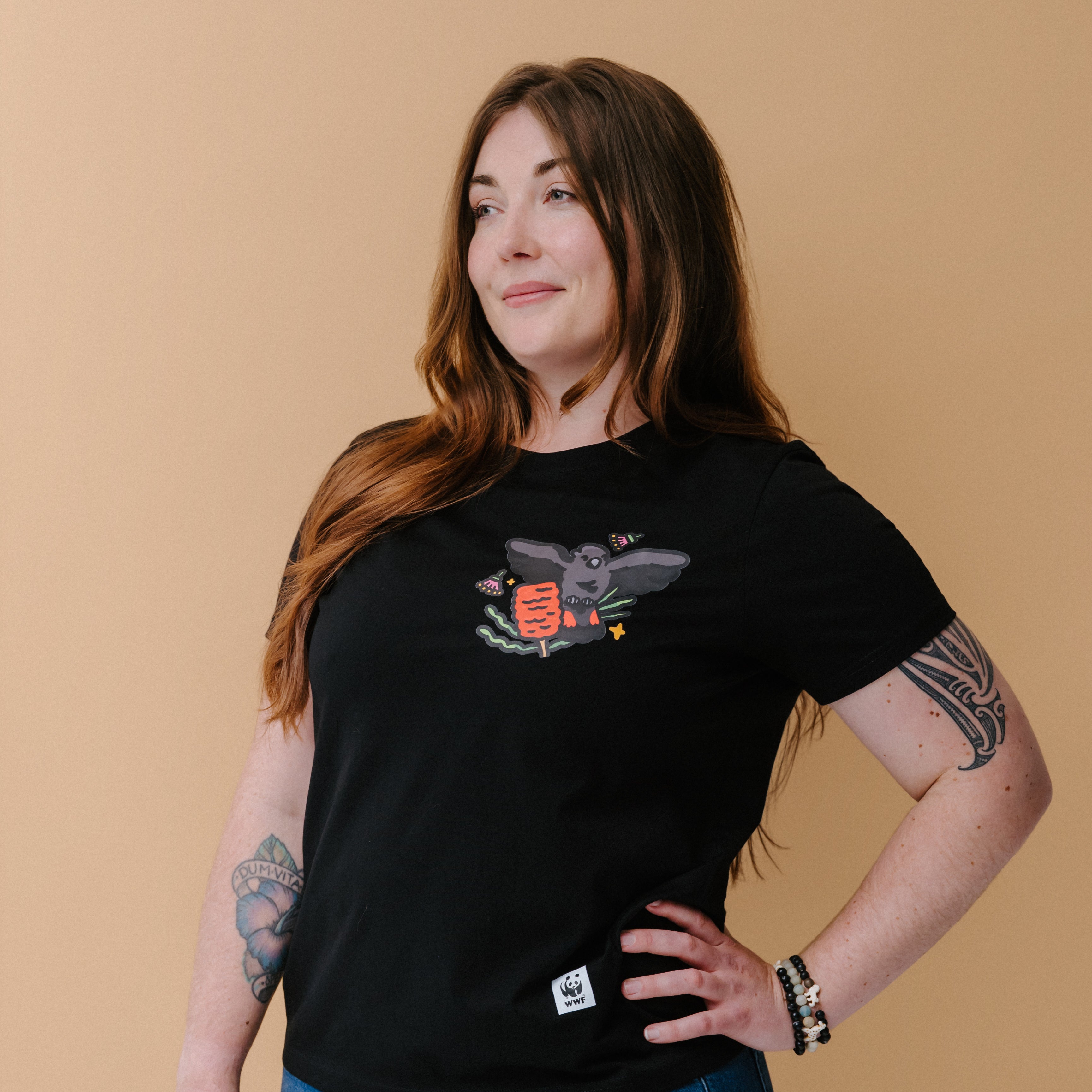 A woman with long brown hair and tattoos wearing a black graphic t-shirt with an illustrated glossy black cockatoo, posing against a solid beige background.