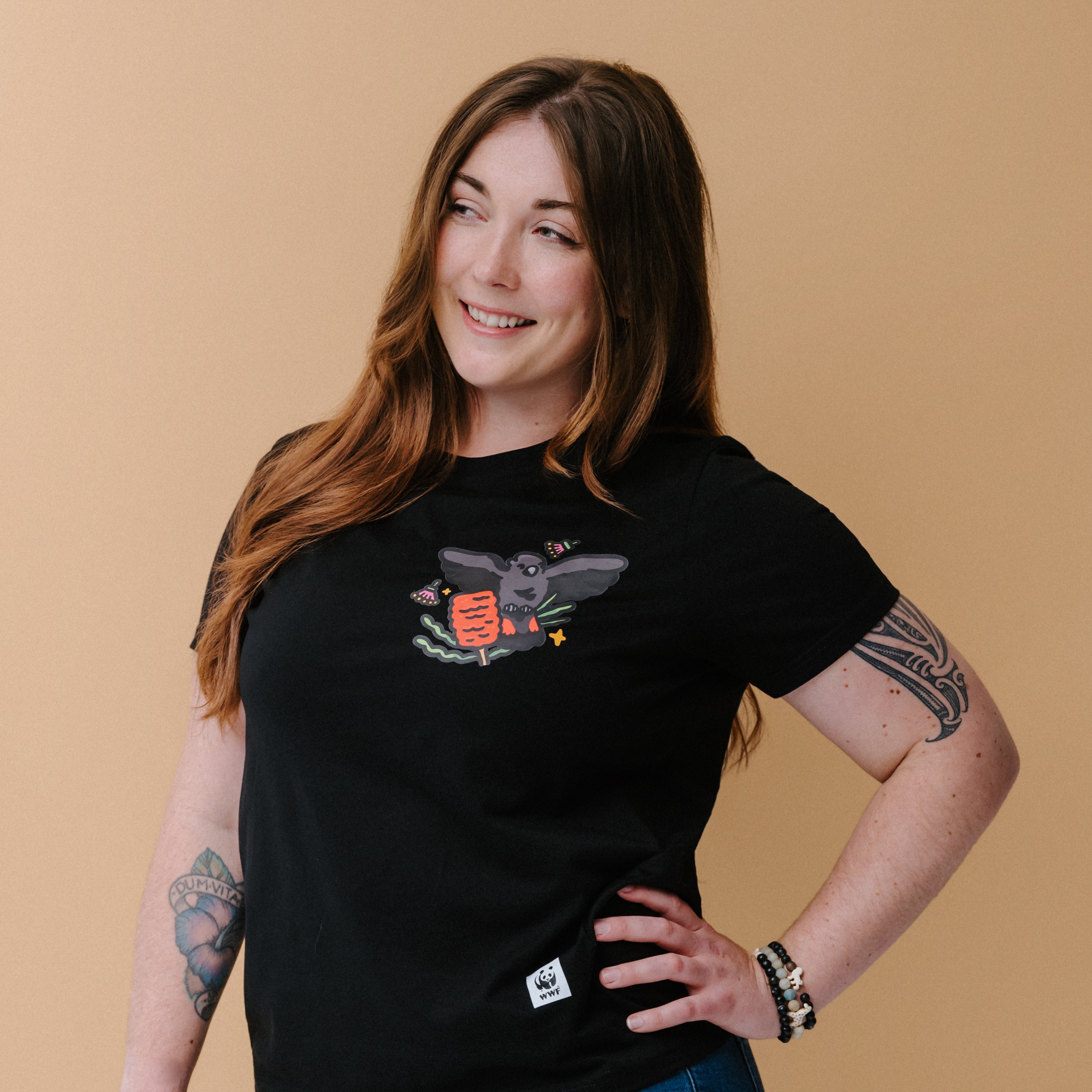 A woman with long brown hair and tattoos wearing a black graphic t-shirt with an illustrated glossy black cockatoo, posing against a solid beige background.