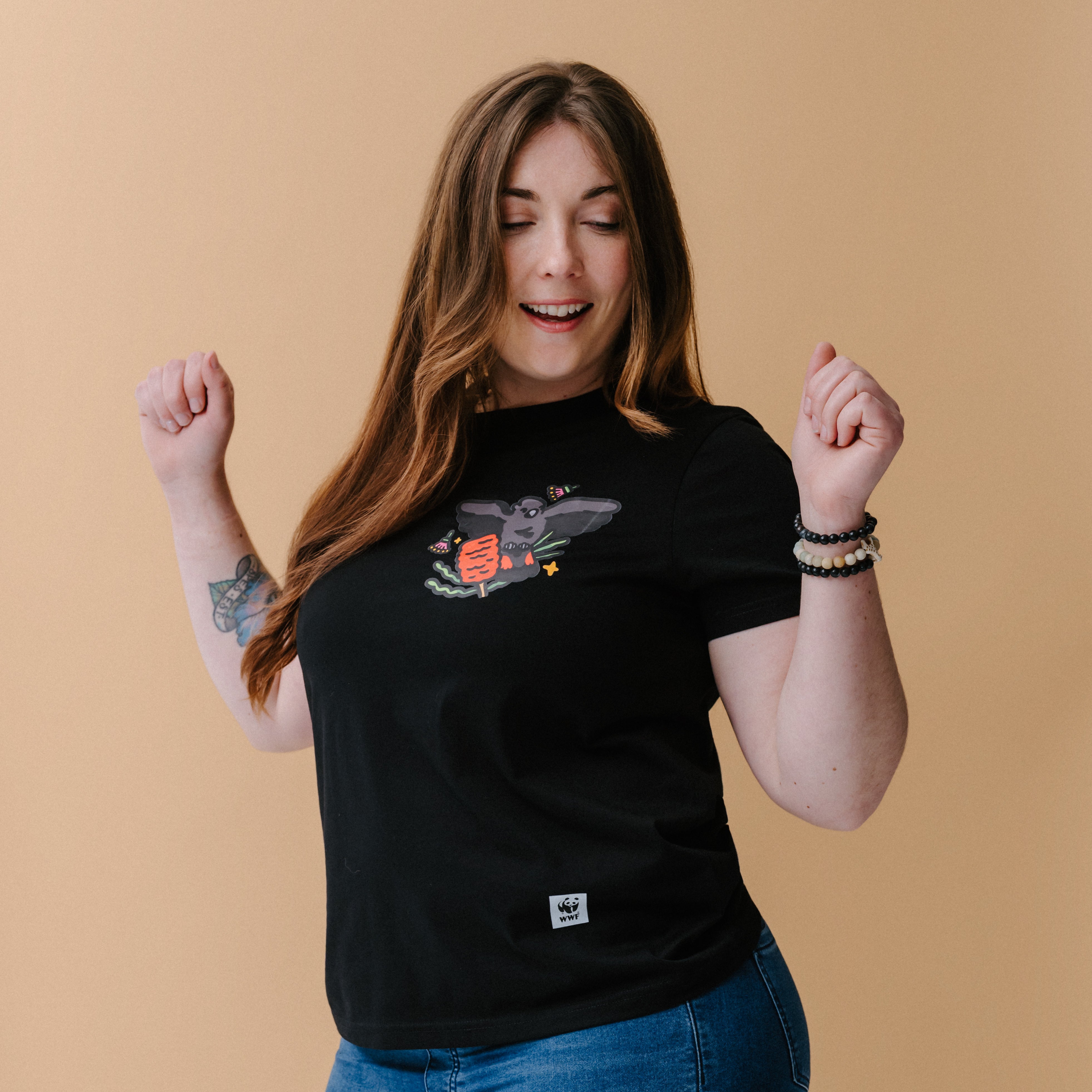 A woman with long brown hair and tattoos wearing a black graphic t-shirt with an illustrated glossy black cockatoo, posing against a solid beige background.