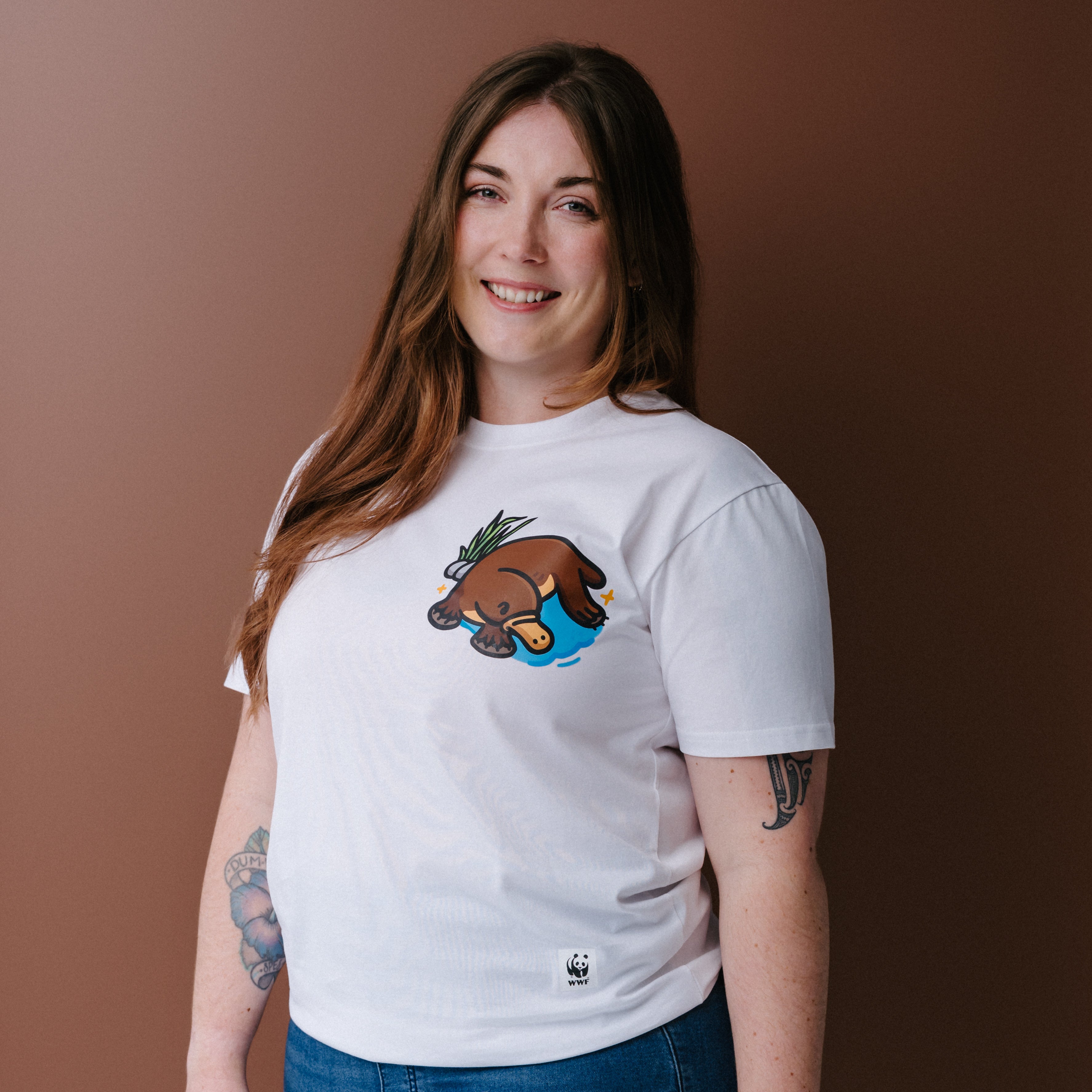 A woman with long brown hair wearing a white crewneck t-shirt with an illustrated platypus graphic on the chest, posed against a solid brown background.
