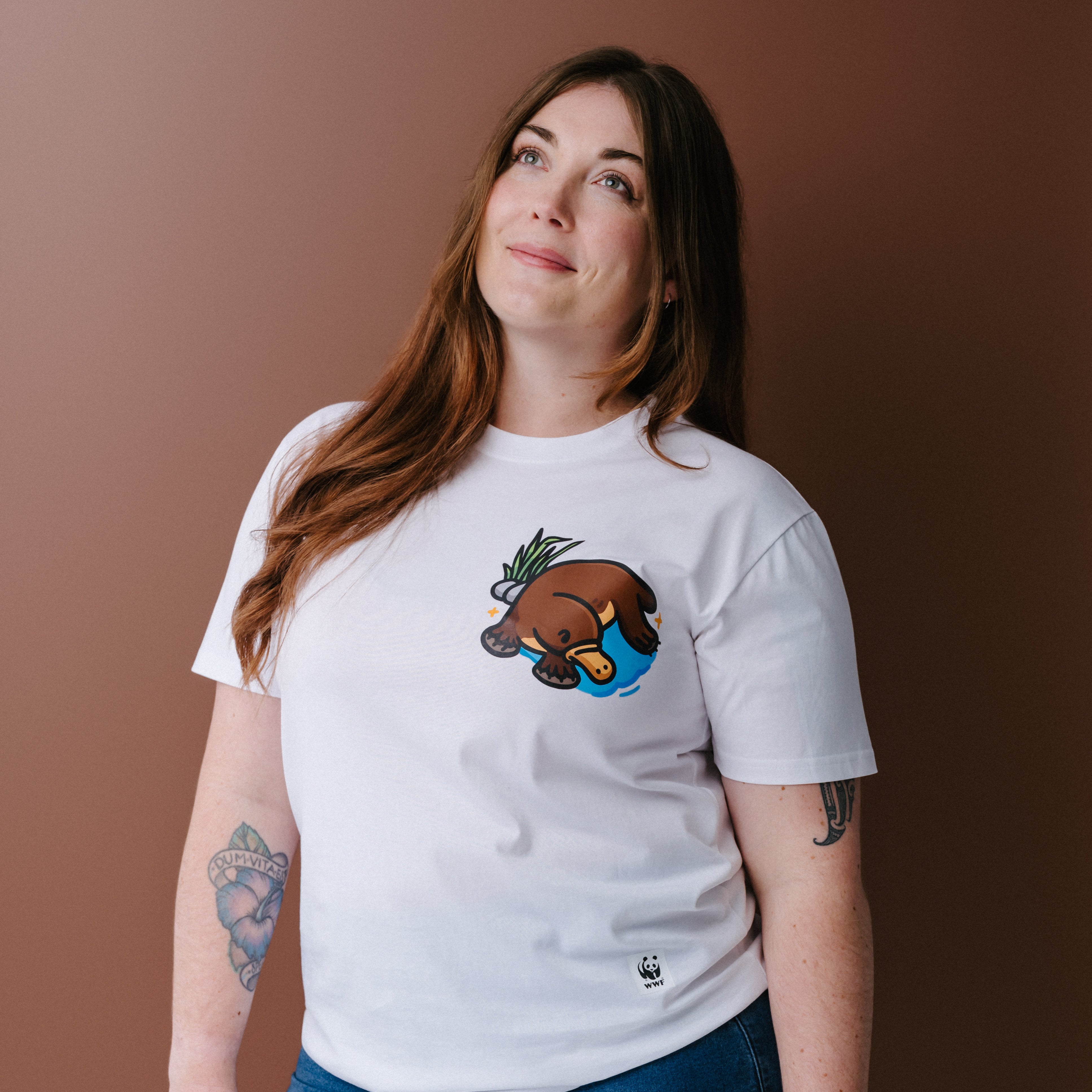 A woman with long brown hair wearing a white crewneck t-shirt with an illustrated platypus graphic on the chest, posed against a solid brown background.