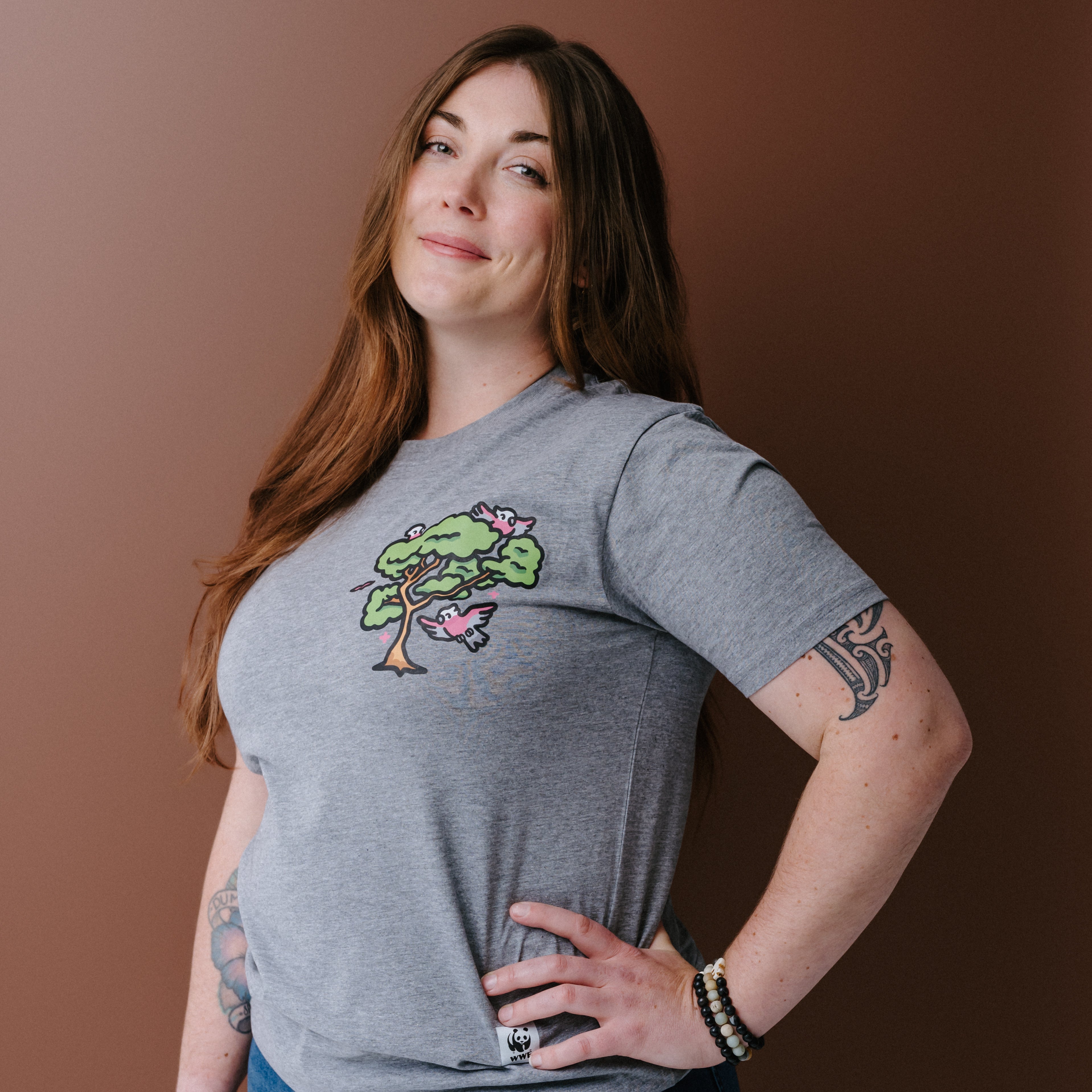 A woman with long brown hair wearing a heather gray t-shirt with a colorful tree and galas illustration on the left chest, posing against a solid brown background.