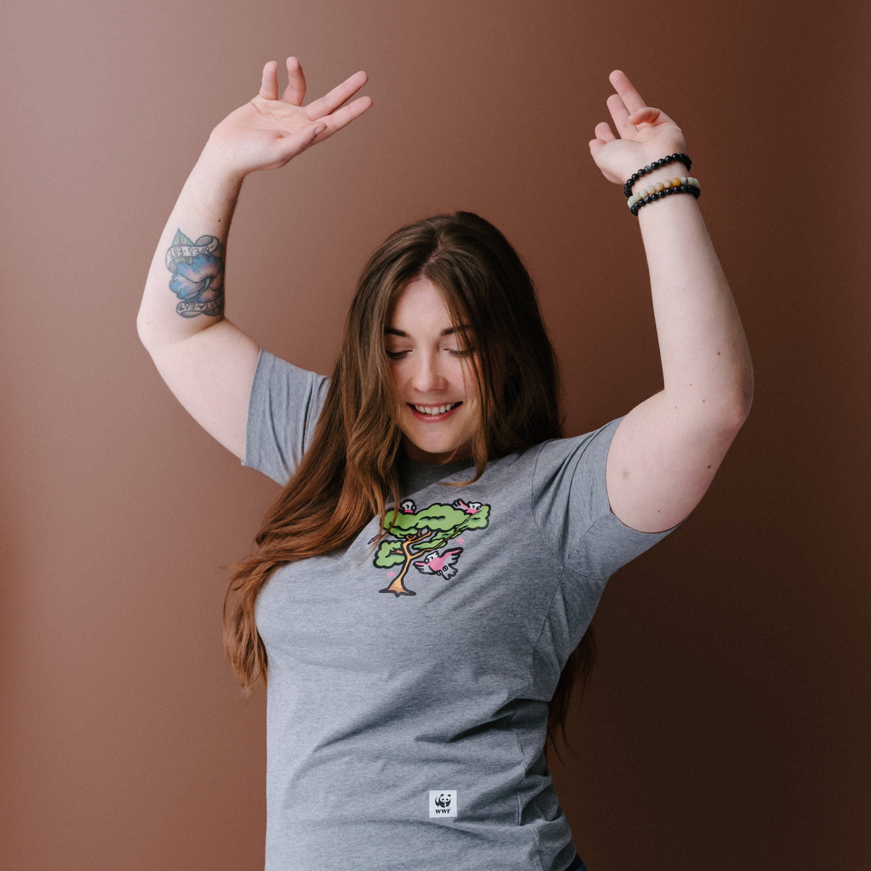 A woman with long brown hair wearing a heather gray t-shirt with a colorful tree and galas illustration on the left chest, posing against a solid brown background.