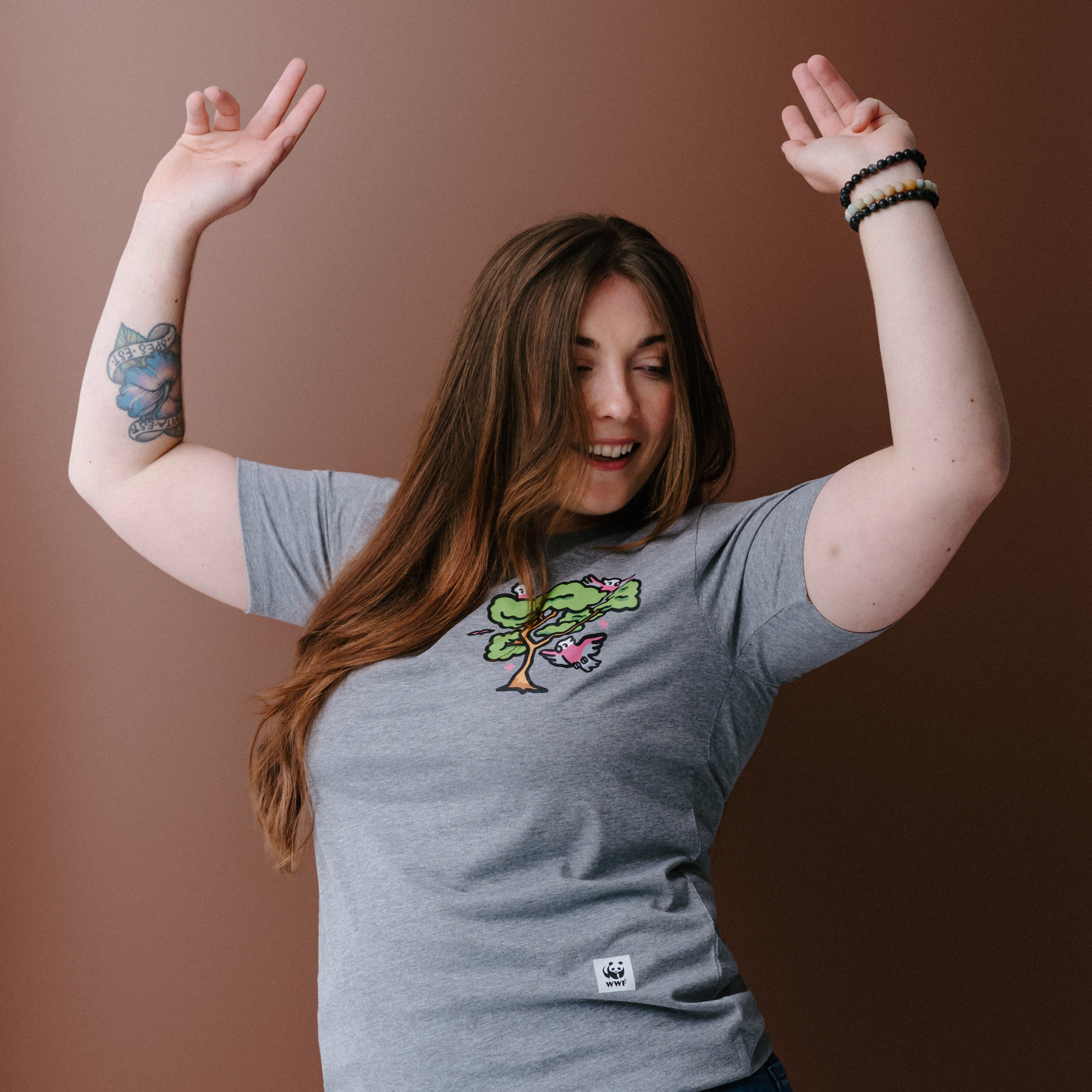 A woman with long brown hair wearing a heather gray t-shirt with a colorful tree and galas illustration on the left chest, posing against a solid brown background.