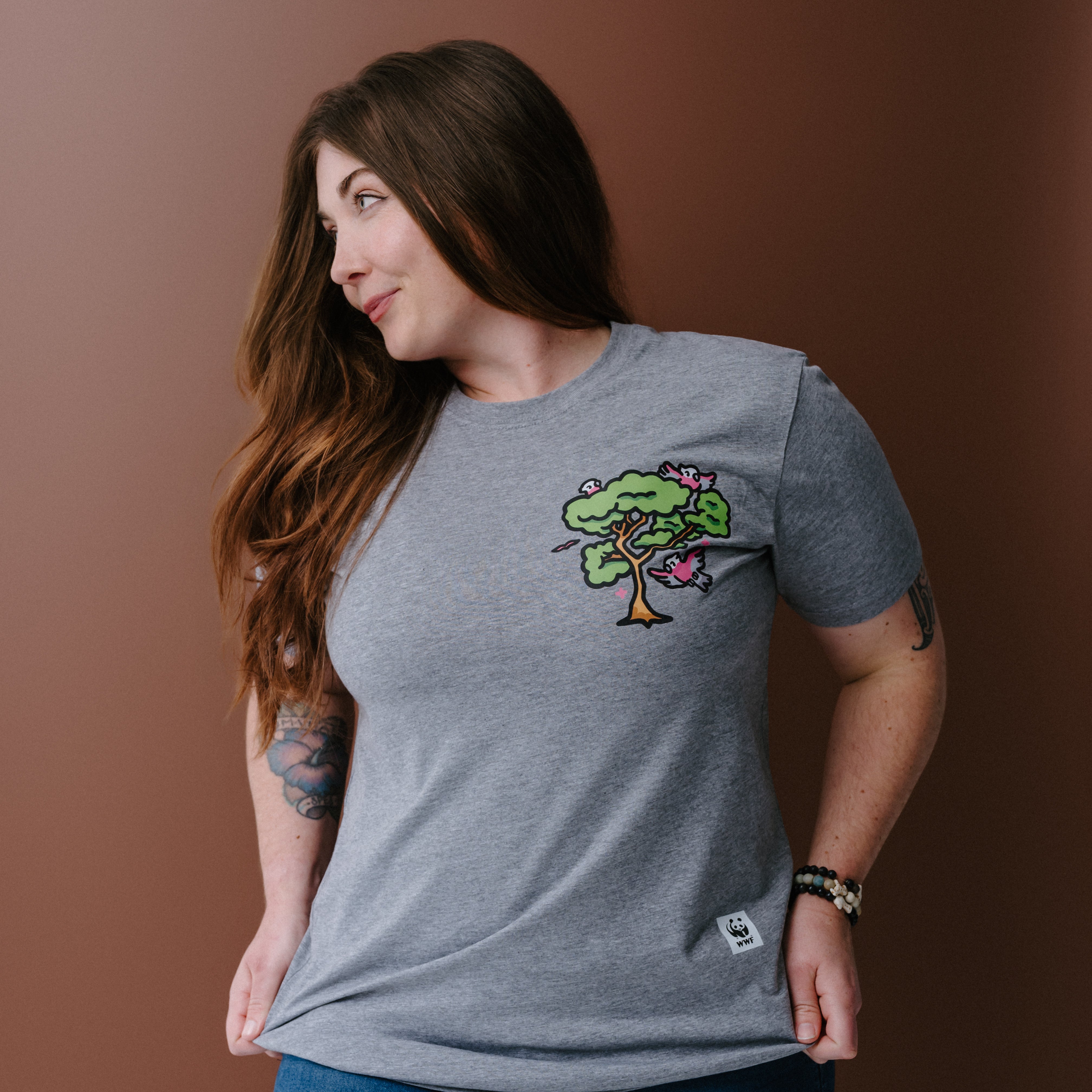 A woman with long brown hair wearing a heather gray t-shirt with a colorful tree and galas illustration on the left chest, posing against a solid brown background.
