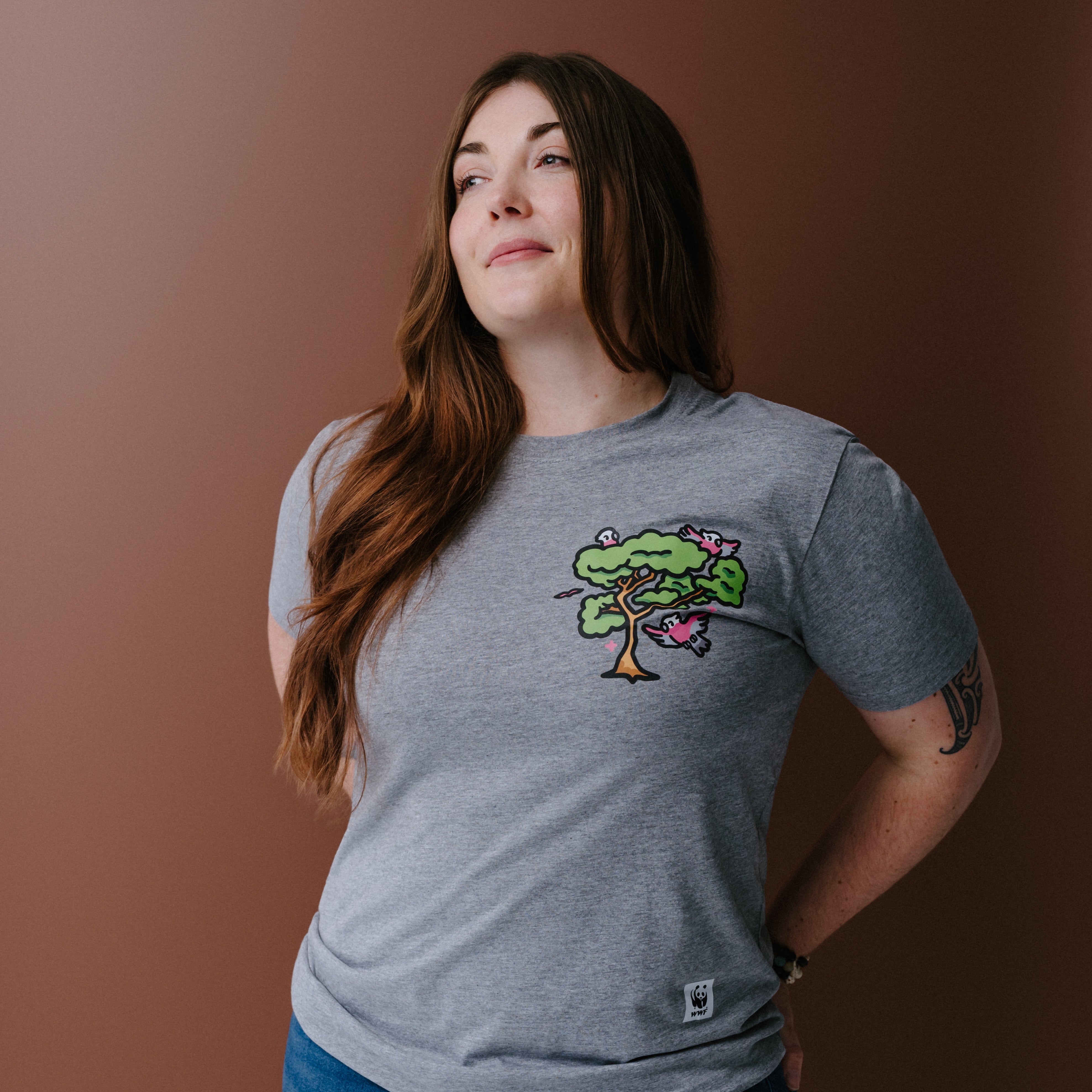A woman with long brown hair wearing a heather gray t-shirt with a colorful tree and galas illustration on the left chest, posing against a solid brown background.